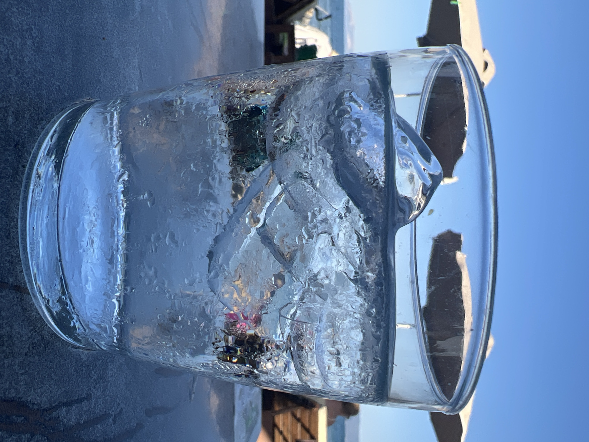 A close-up, low-angle shot focuses on a glass filled with ice and water. Condensation beads on the exterior of the glass, which sits on a dark, textured surface. In the background, out of focus, are beach umbrellas, lounge chairs, and a body of water, suggesting a beach or seaside location. The sky is clear and blue. The location is Eretria, Eretria Municipal Unit, Greece.