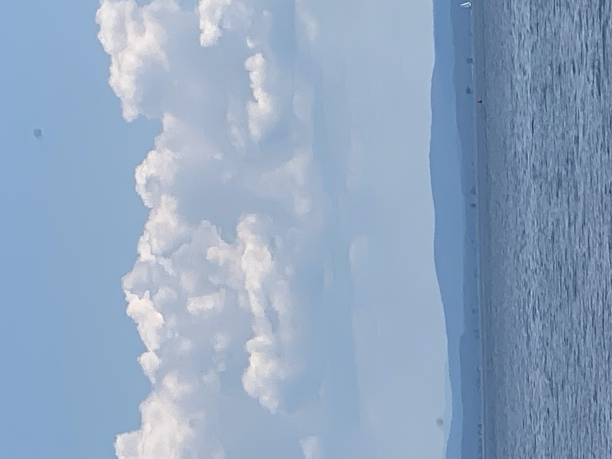 The image captures a scenic view of a body of water next to a coastline, with a bright blue sky filled with fluffy white clouds overhead. The water has a textured appearance, suggesting gentle waves or ripples. In the distance, faint landmasses are visible, appearing as soft blue layers that recede into the horizon, hinting at a hazy atmosphere or the curvature of the Earth.
On the distant shoreline, small structures or trees are subtly discernible. A tiny red object, possibly a marker or a buoy, is visible on the water near the coast. There are also what appear to be small white specks in the distance, which could be sailboats or distant objects. One such speck, a small dark square, is in the upper left portion of the sky. The overall scene suggests a peaceful day, likely during daylight hours given the bright sky and clouds. The location is described as the Town of Peru, United States of America, though no specific landmarks within the town are identifiable from this image. The weather appears to be clear and pleasant, conducive to outdoor activities.