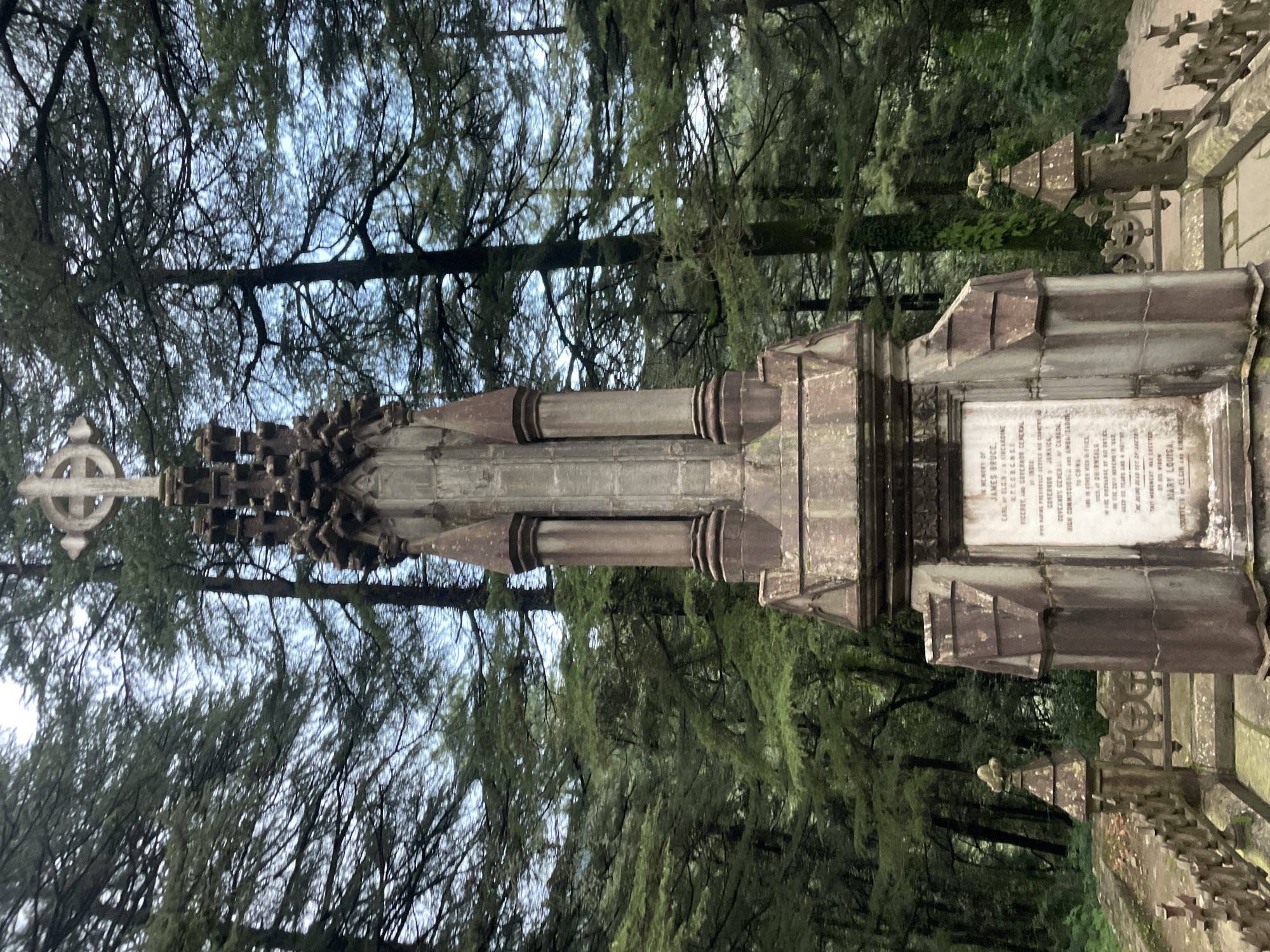 This is a full-frame, eye-level, vertical shot of a weathered stone monument in a forested area. The monument is tall and ornate, featuring a cross at the top, intricate stonework, and a plaque with inscriptions. The plaque appears to list names and titles, likely a memorial. Flanking the main monument are smaller, similar stone structures and a decorative stone railing. The background is dominated by tall, dark green coniferous trees, suggesting a cool, possibly mountainous climate. The sky peeks through the branches, appearing overcast or a pale blue. In the middle-right background, a dark-colored animal, possibly a dog, is lying down. The overall scene evokes a sense of solemnity and history, situated within a natural, secluded environment. The location is identified as Dharamkot, India.