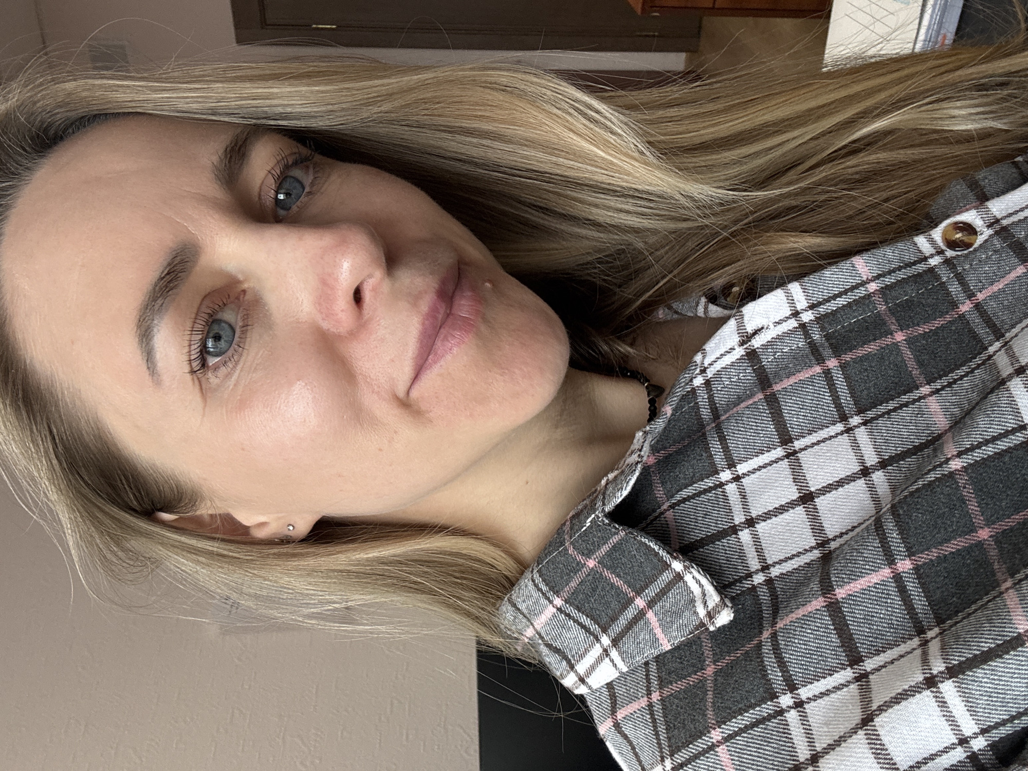 The image displays a close-up, portrait-style photograph of a woman, with the shot taken from an upward angle and rotated approximately 90 degrees clockwise. The woman has long, straight blonde or light brown hair, parted on her left side, flowing over her shoulders. She possesses striking blue eyes with long eyelashes and neatly groomed dark eyebrows. Her skin appears fair, with a few subtle lines visible on her forehead. A small, light-colored blemish is noticeable on her upper lip. She wears a small silver stud piercing on her left ear's tragus. Her expression is calm and neutral, with a gentle, slight smile.
She is dressed in a plaid button-down shirt featuring a pattern of dark grey, white, and pink squares. A dark, thin strap, possibly part of a necklace or garment, is partially visible around her neck. The background is simple, comprising a plain light beige or tan wall surface on one side and a darker brown wooden element, possibly furniture or a door frame, on the other. The lighting in the image is soft and natural, suggesting an indoor setting during the daytime. No specific location cues, such as landmarks or signage, are visible in the image to indicate Chaikovsky, Russia, nor is there any discernible text. The photograph captures an intimate and casual moment, focusing primarily on the woman's face and upper body.