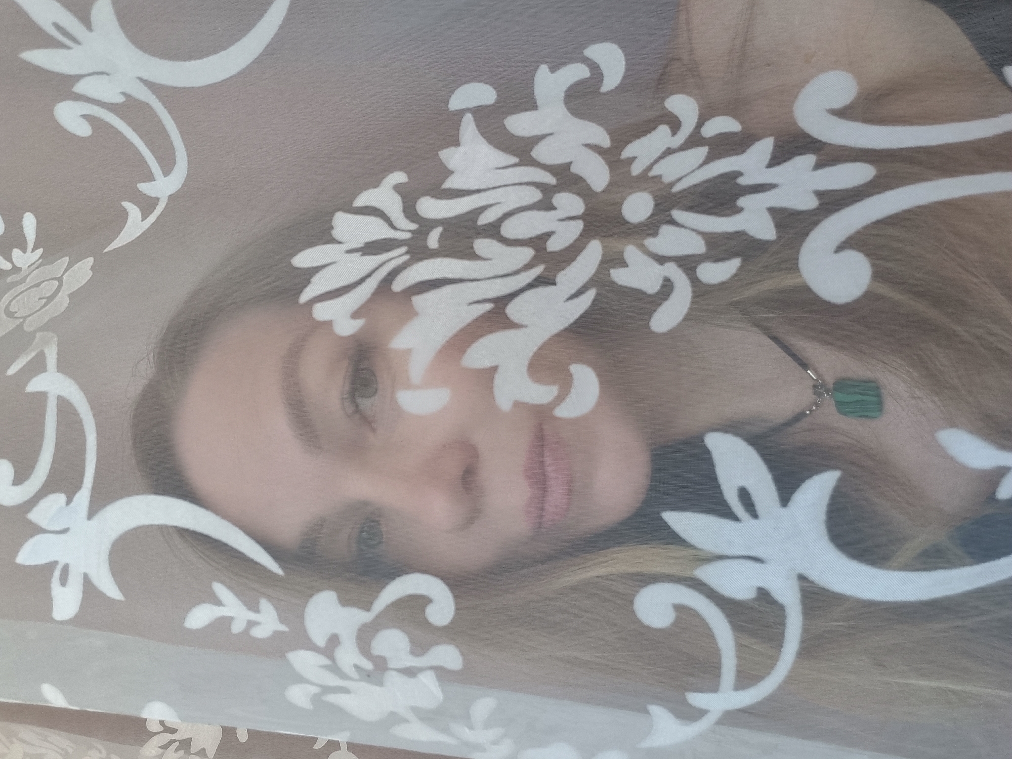 The image shows a woman's face partially obscured by a patterned fabric. The fabric features a large, repeating white floral or damask pattern. The woman has fair skin, blonde hair, and light-colored eyes. She wears a necklace with a square, green pendant.
There is no evidence in the image to identify the location as Chaikovsky, Russia.