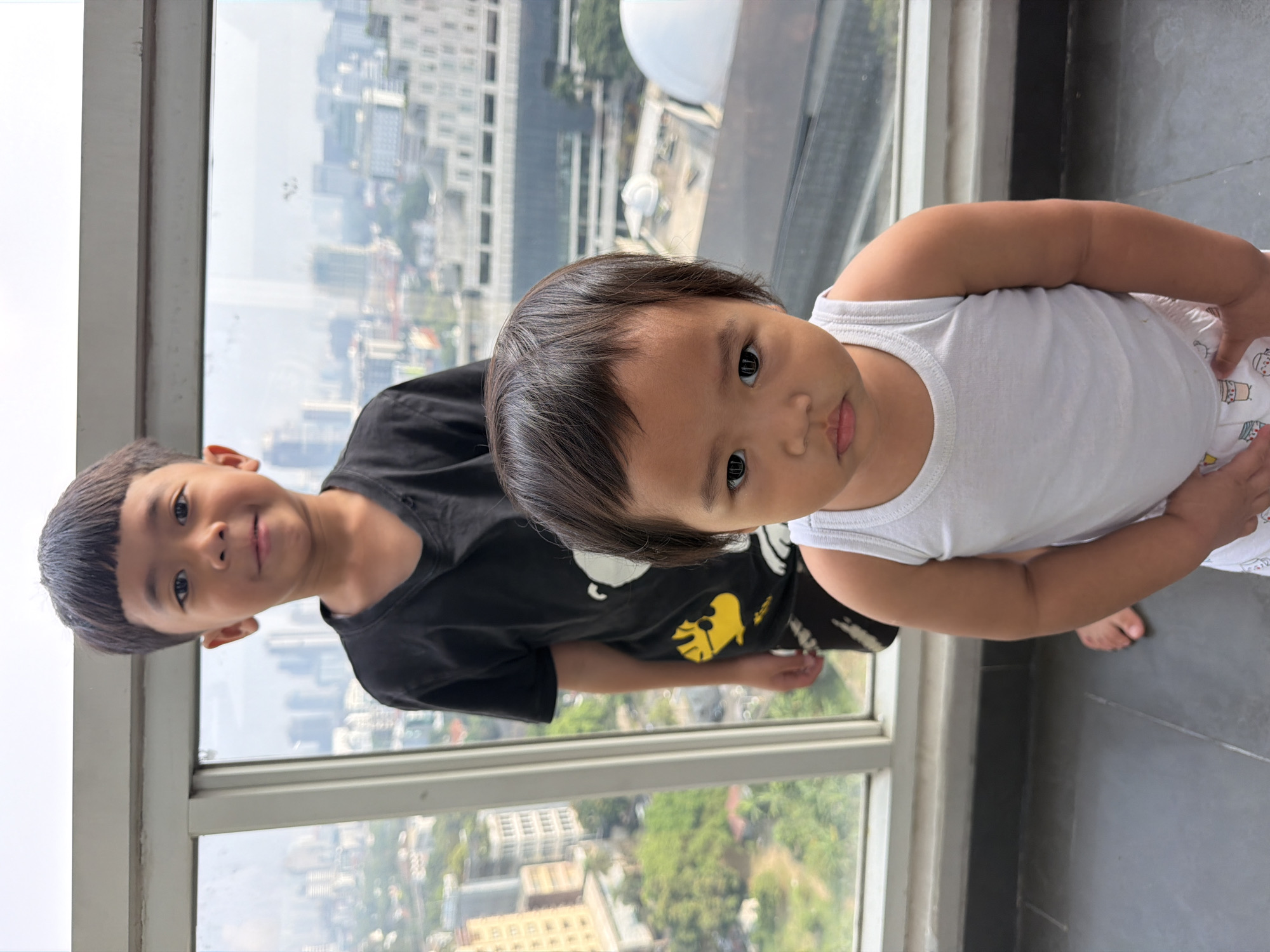 Two young boys stand in front of a window with a city view. The boy in the foreground, closer to the camera, is wearing a white tank top and printed shorts. He has dark hair and is looking directly at the camera with a neutral expression. The boy in the background, taller and behind the first boy, is wearing a black t-shirt with a graphic print and patterned shorts. He also has dark hair and is looking towards the camera with a slight smile. The background reveals a cityscape with buildings and greenery, suggesting an urban environment. The location is identified as Central Jakarta, Indonesia.