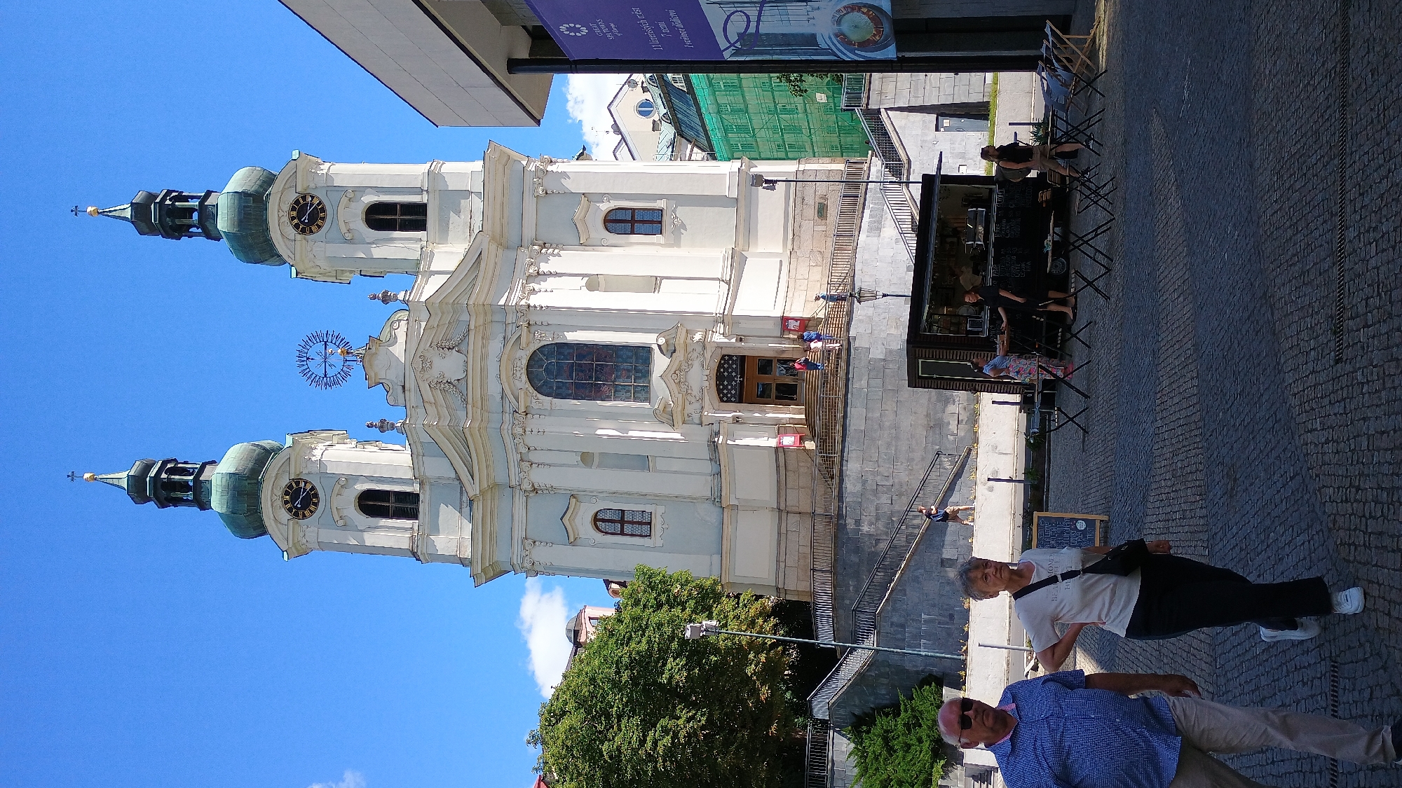 The image depicts a daytime scene in the city of Karlovy Vary, Czechia, likely near the St. Mary Magdalene Church. In the foreground, an older couple walks across a cobblestone plaza. The man, on the left, wears a blue checkered shirt and light-colored pants. The woman, on the right, is dressed in a light-colored t-shirt with dark pants and a crossbody bag. Behind them, a food truck is parked, with several people standing at the counter. Some tables and chairs are set up in front of the truck.
The background is dominated by a large, ornate white church with two prominent towers topped with green domes. One tower features a clock face. A wide staircase leads up to the church entrance, and several people can be seen ascending or descending it. Lush green trees are visible to the left of the church, and modern buildings with scaffolding and green sheeting are present on the right side of the image. The sky is a clear blue with a few scattered white clouds. The overall atmosphere appears to be a sunny, pleasant day in a European city.