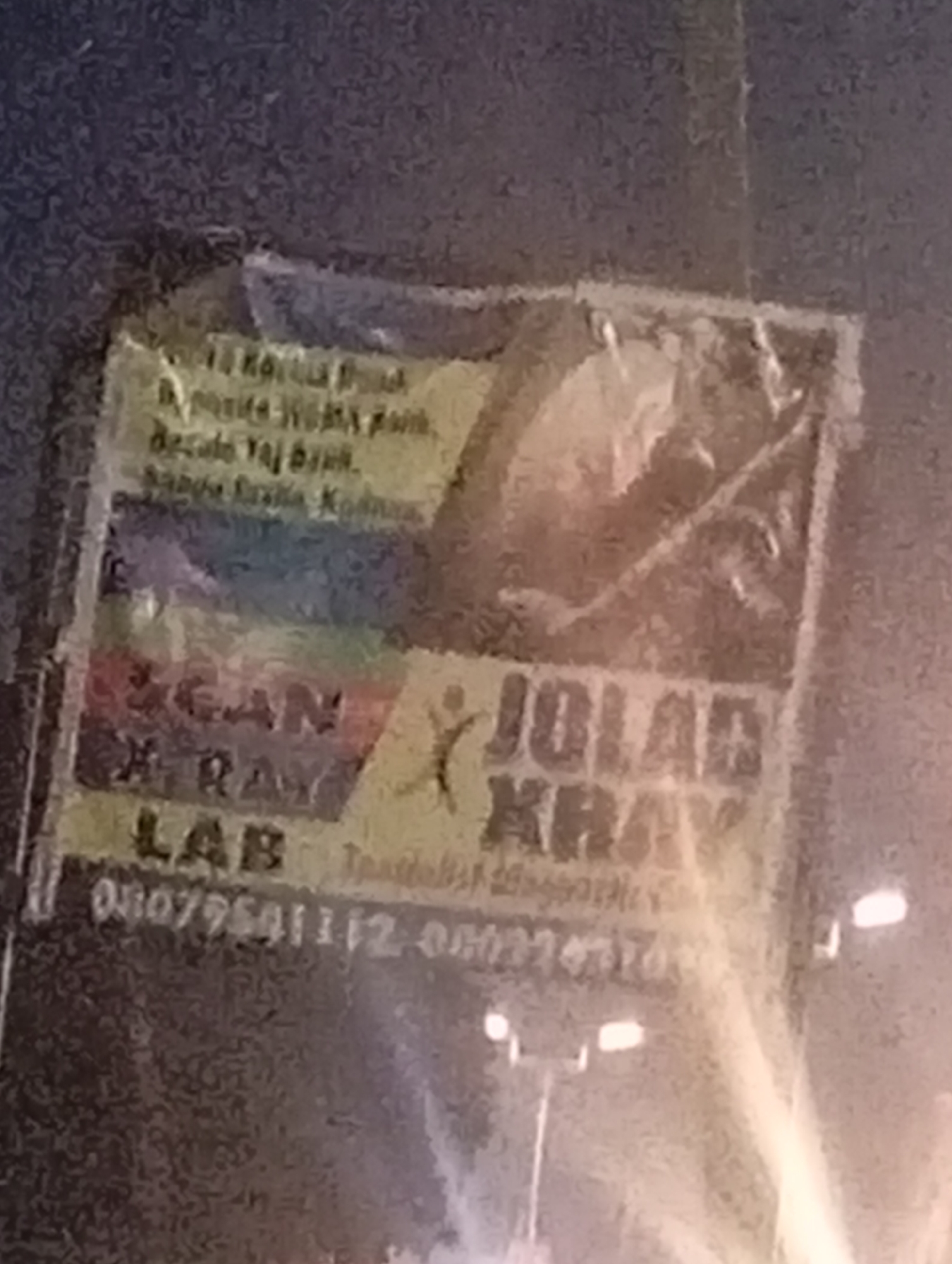 The image shows a close-up of a faded, slightly torn poster or banner attached to a dark, textured surface, possibly a wall or a building exterior. The poster appears to be an advertisement for a medical service. Text on the poster is partially visible and difficult to read due to the image's low resolution and blurriness.
Several key phrases and words can be deciphered, though with some uncertainty. At the top, there's text that seems to be an address or location, possibly including "Sabon Tasha, Nigeria." Below this, the words "SCAN," "X RAY," and "LAB" are clearly visible, indicating these are the services offered. Another prominent part of the text reads "JOLAR XRAY."
There is a small graphic of a stylized human figure with outstretched arms on the left side of the "JOLAR XRAY" text. Below the service listings, a phone number is partially visible, starting with "080775..." and followed by more digits that are unreadable.
The poster has a rainbow-colored band across the middle, and the background of the poster features a faint image of a person's face or a silhouette. The overall condition of the poster suggests it has been exposed to the elements. The surrounding environment is dark, with some out-of-focus lights visible in the background, suggesting the photo was taken at night or in a dimly lit area.