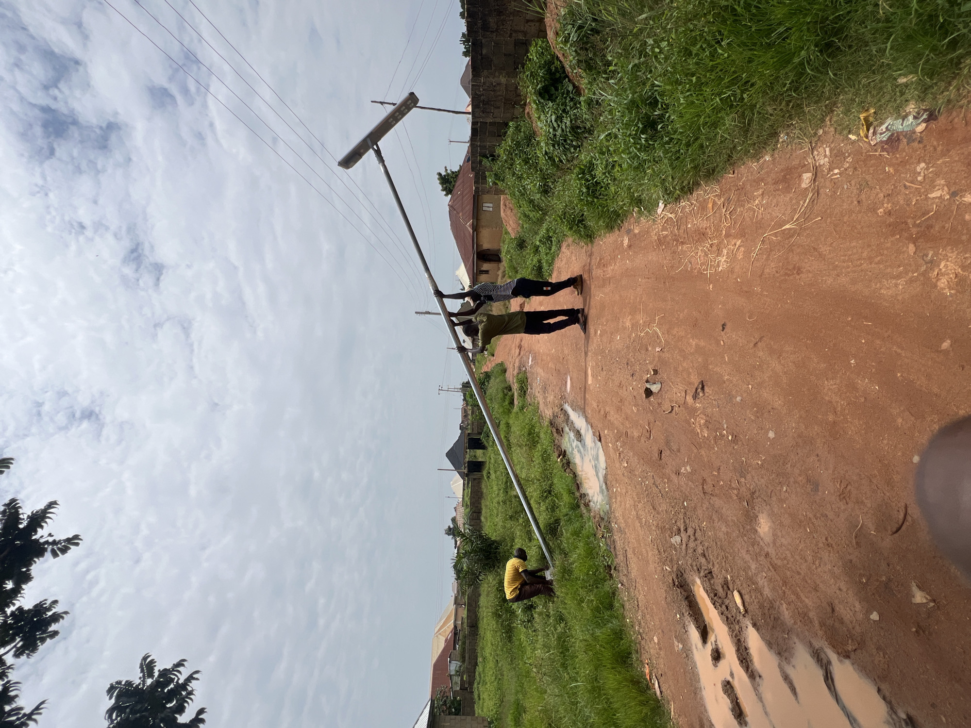 Two young men are carrying a large, light-colored metal pole with a street light attached to one end. They are walking on a dirt road in what appears to be a residential area. One man is wearing a dark green t-shirt and dark pants, while the other is wearing a striped shirt and dark pants. A third person, wearing a yellow and brown striped shirt, is bent over to the left of the road, possibly working on something on the ground. The road is muddy in places with large puddles. Houses with red tiled roofs and brick walls are visible in the background, along with green vegetation and trees. The sky is cloudy. The scene is likely in Jos, Nigeria, given the visual cues of the environment and architecture.
