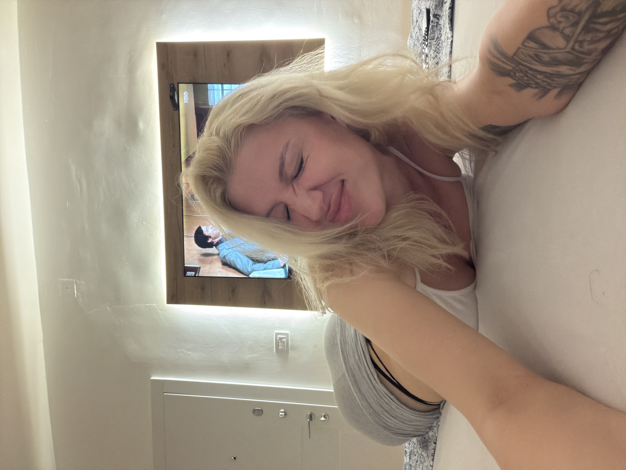 A medium shot shows a blonde woman lying on a white pillow in a bedroom. She is wearing a white tank top and a grey striped shirt. The woman has a tattoo on her left arm. Behind her is a television set mounted on the wall, with a light panel behind it. The scene appears to be set in a hotel room, and the environment suggests the city of Belgrade, Serbia.
