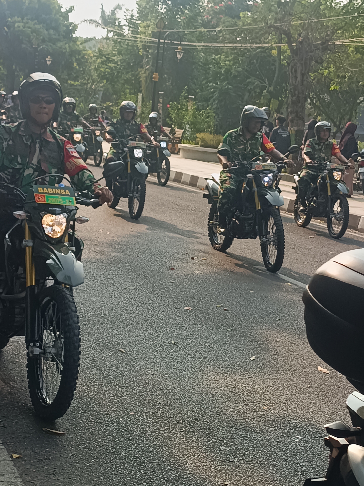 The image captures a group of individuals, likely members of the Indonesian military, riding motorcycles in a procession. They are dressed in camouflage uniforms and helmets. The motorcycles are of a dual-sport type, with knobby tires suggesting they are capable of off-road use. One motorcycle in the foreground displays a license plate with the text "BABINSA 29075-10". The procession appears to be on a paved road, with trees and some buildings visible in the background. The scene is illuminated by natural sunlight, casting shadows on the road. The presence of the "BABINSA" designation on the license plate indicates this event is likely related to a community policing or outreach program within the Indonesian National Armed Forces, common in rural and urban areas across Indonesia, including Sleman, Yogyakarta.