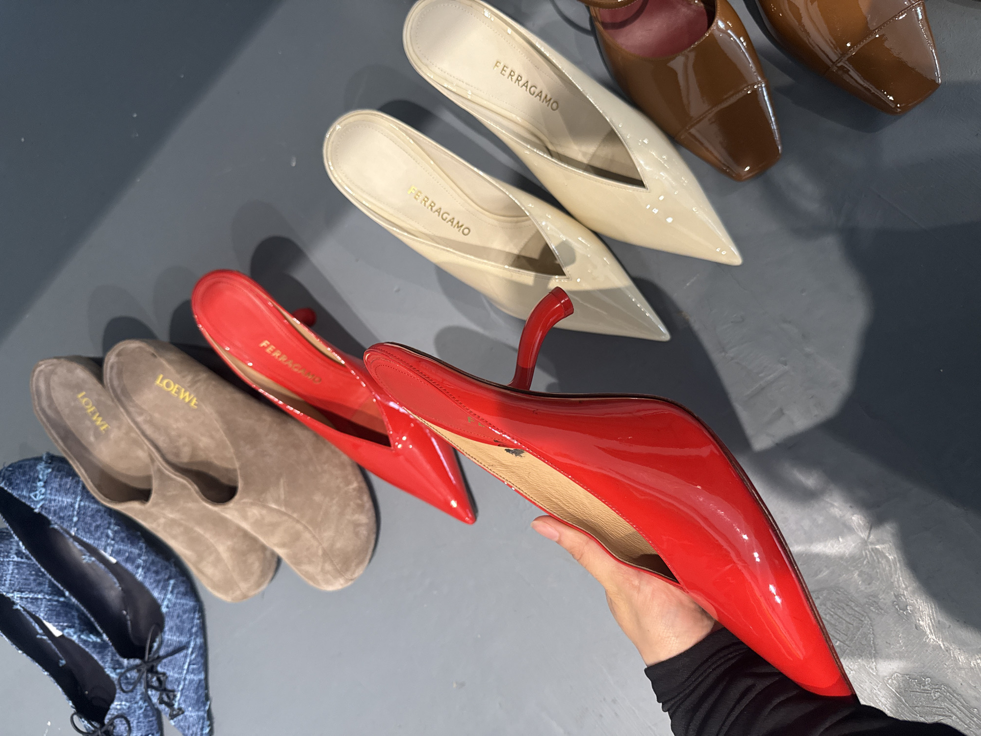 The image shows a selection of women's shoes arranged on a gray surface. Several brands are visible, including Ferragamo and Loewe. The color palette ranges from neutral tones in the Ferragamo mules and Loewe slippers to bright red in the pointed-toe heels. One red shoe is being held by a hand, while the others are displayed on the surface. The background is a plain gray, suggesting a studio or showroom setting. The overall impression is one of a curated collection of designer footwear. There is no information in the image to suggest this scene is in London or the United Kingdom.