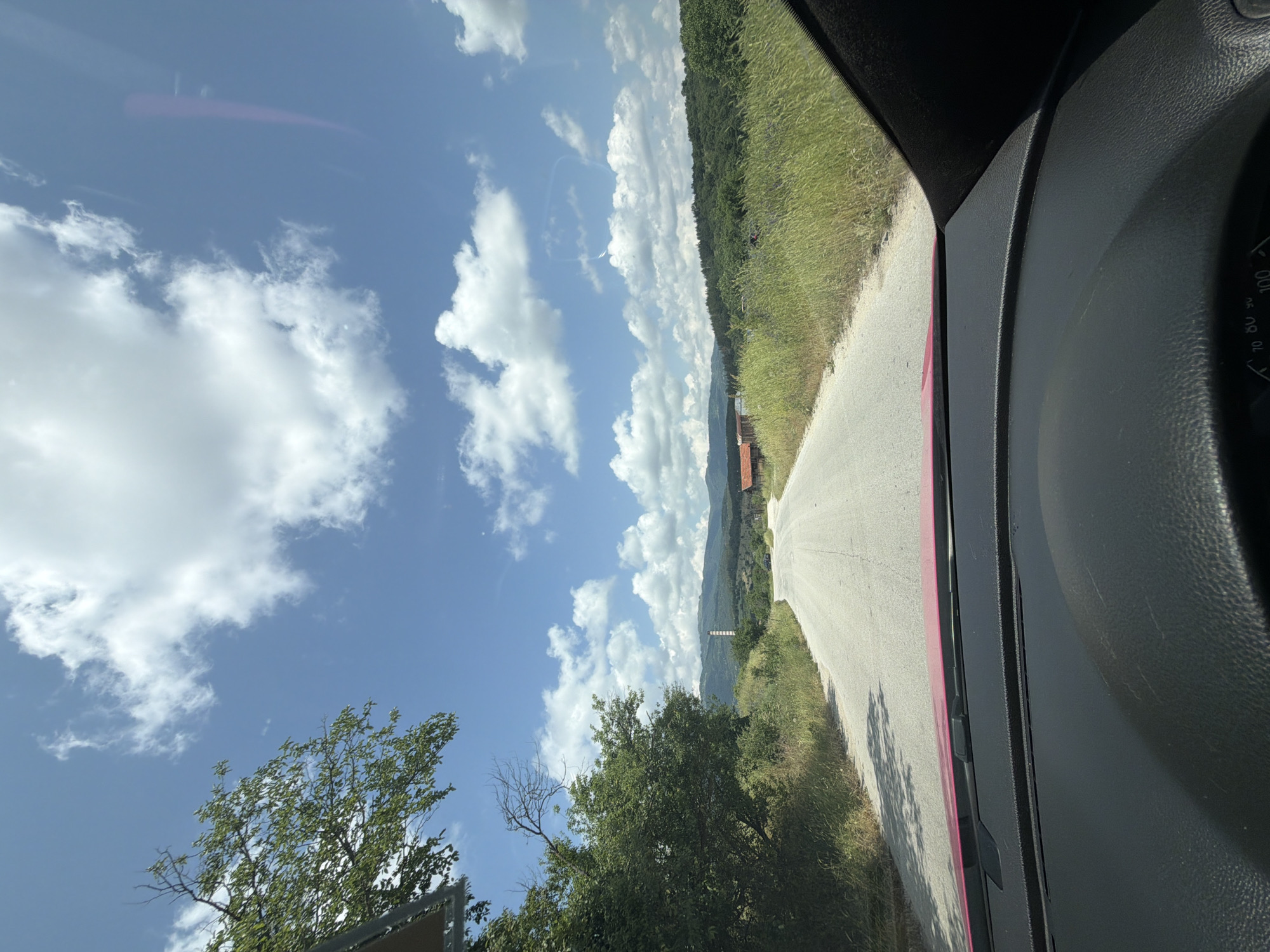 The original image is captured from inside a vehicle, looking out at a scenic, sunny landscape, and is presented rotated 90 degrees clockwise. When viewed upright, the scene depicts a clear blue sky with scattered white cumulus clouds dominating the upper portion of the frame.
In the foreground, the right side of the image shows the dark interior of a vehicle, including a textured dashboard, part of the windshield frame, and a partial view of a gauge cluster displaying numbers like "70, 80, 90, 100," suggesting a speedometer or similar dial. A bright red reflection or component is visible along the edge of the windshield, indicating a possible red vehicle.
To the left, a paved road stretches into the distance, flanked by green grassy verges and a line of trees and dense foliage. The road appears to curve slightly to the right. Beyond the road, the mid-ground features more lush green trees and rolling hills or mountains. A few distant buildings are visible, including one with a distinctive red-tiled roof, and a tall, slender white structure, possibly a chimney or communication tower, stands out against the green backdrop. A large, leafy green tree with some bare branches occupies the lower-left corner, close to the viewer.
The image conveys a bright, clear day, likely midday or early afternoon, characterized by strong sunlight and visible shadows. The setting suggests a drive through a rural or semi-rural area, possibly around Strelcha, Bulgaria, given the location context provided. The overall mood is one of travel and appreciation for nature under a vibrant sky. No other specific text is clearly legible apart from the gauge numbers.