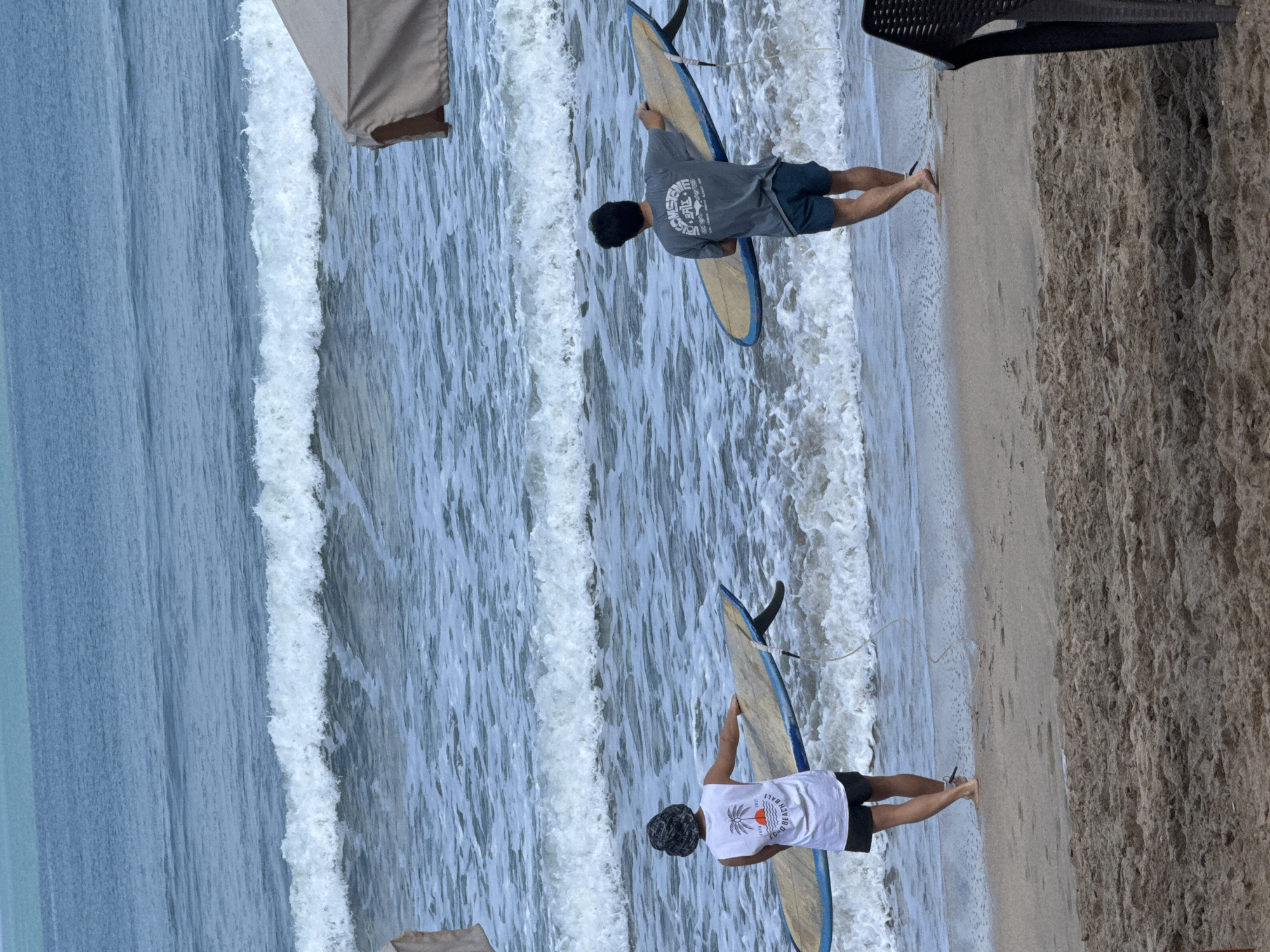 Two individuals are walking into the ocean on a sandy beach in Kuta, Indonesia, each carrying a surfboard. The person on the left, wearing a white tank top and black shorts, has a bucket hat on. The person on the right is wearing a grey t-shirt and blue shorts. The ocean has moderate waves, and there are parts of a beach umbrella visible in the upper right portion of the image. The sand appears to be dark and damp near the water's edge.