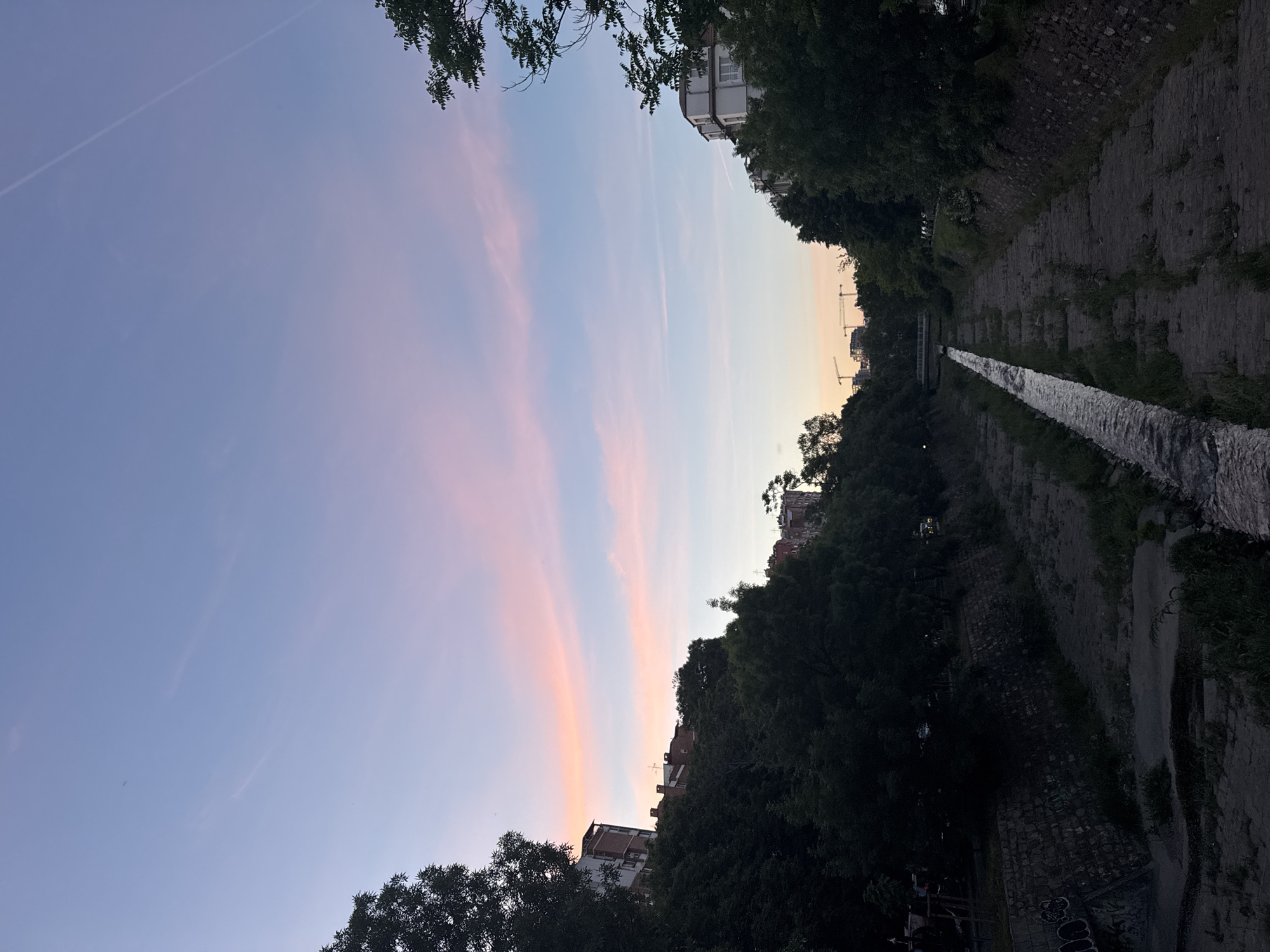 Captured at dusk in Sofia, Bulgaria, the photograph is oriented vertically and shows a narrow, concrete-lined canal stretching into the distance, filled with flowing water. On either side, dense greenery lines the banks, with buildings intermittently visible above the foliage. The sky above displays a gradient of color, transitioning from a light blue at the top left to a soft orange near the horizon. A few wispy contrails are visible in the sky. Some graffiti appears on the concrete in the bottom right corner of the frame. Construction cranes appear to be visible in the far distance, on the horizon.