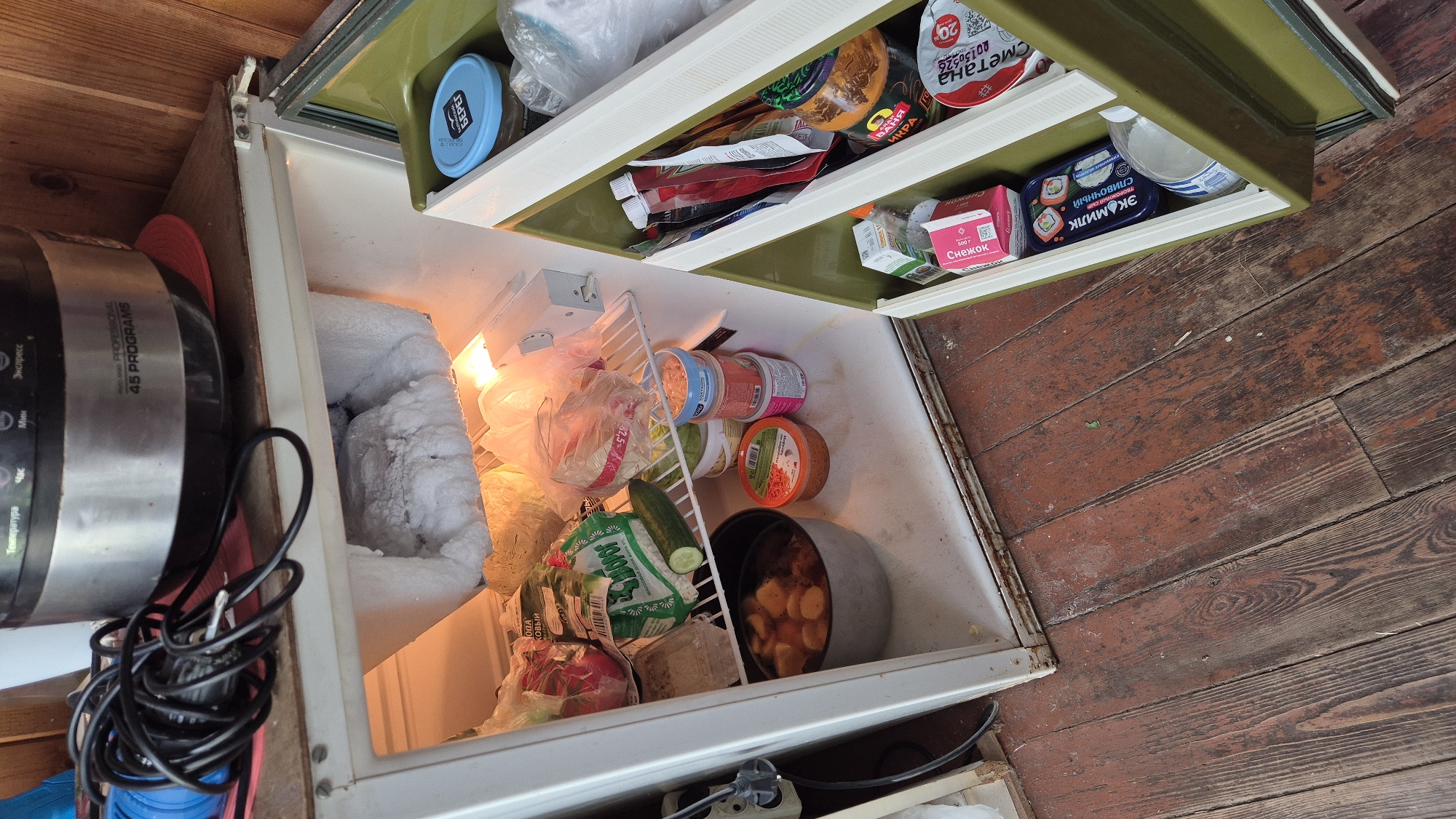 This is a medium shot, eye-level, exterior photograph of an open, old, white, two-door refrigerator. The refrigerator is situated on a wooden floor, likely in a rustic or outdoor setting. The upper compartment appears to be a freezer, with significant frost accumulation on the back wall and items below. The main compartment of the refrigerator is filled with various food items. On the top shelf, there are plastic bags containing produce, including what appears to be cabbage and radishes. A cucumber is also visible on this shelf. Below this, there is a large pot containing food, possibly stewed potatoes, and several small containers of what look like spreads or dips. The refrigerator door itself has multiple shelves, stocked with jars, packets, and containers of food, including what appear to be yogurt or sour cream containers, and a jar with a red label that might be jam or sauce. The overall impression is of a functional, albeit somewhat aged, refrigerator containing a variety of food staples. The setting suggests a practical storage solution in an environment where a modern kitchen may not be available. The wooden floor boards are worn and appear to be aged, contributing to the rustic aesthetic. The presence of a multi-cooker on top of the refrigerator indicates it may be placed in a temporary or auxiliary kitchen area. The specific location of Машезеро, Russia, suggests a rural or possibly dacha (summer house) setting.