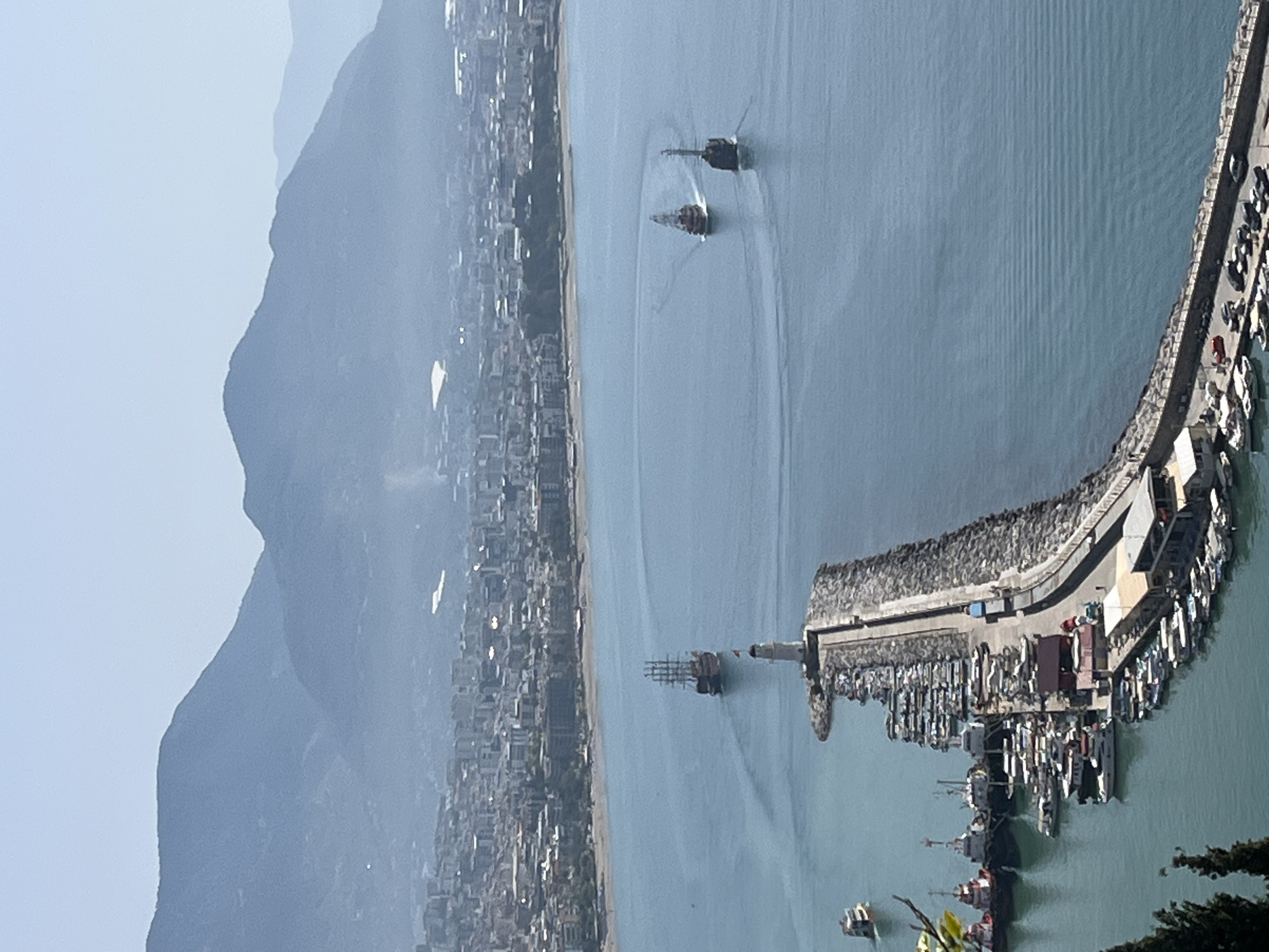 The image presents an elevated view of a coastal scene in Alanya, Turkey. A city sprawl extends along the coastline, backed by mountains. The clear blue water hosts three ships moving away from the shore. A pier, populated with boats, juts into the water from the coastline. The sky is clear and light blue.