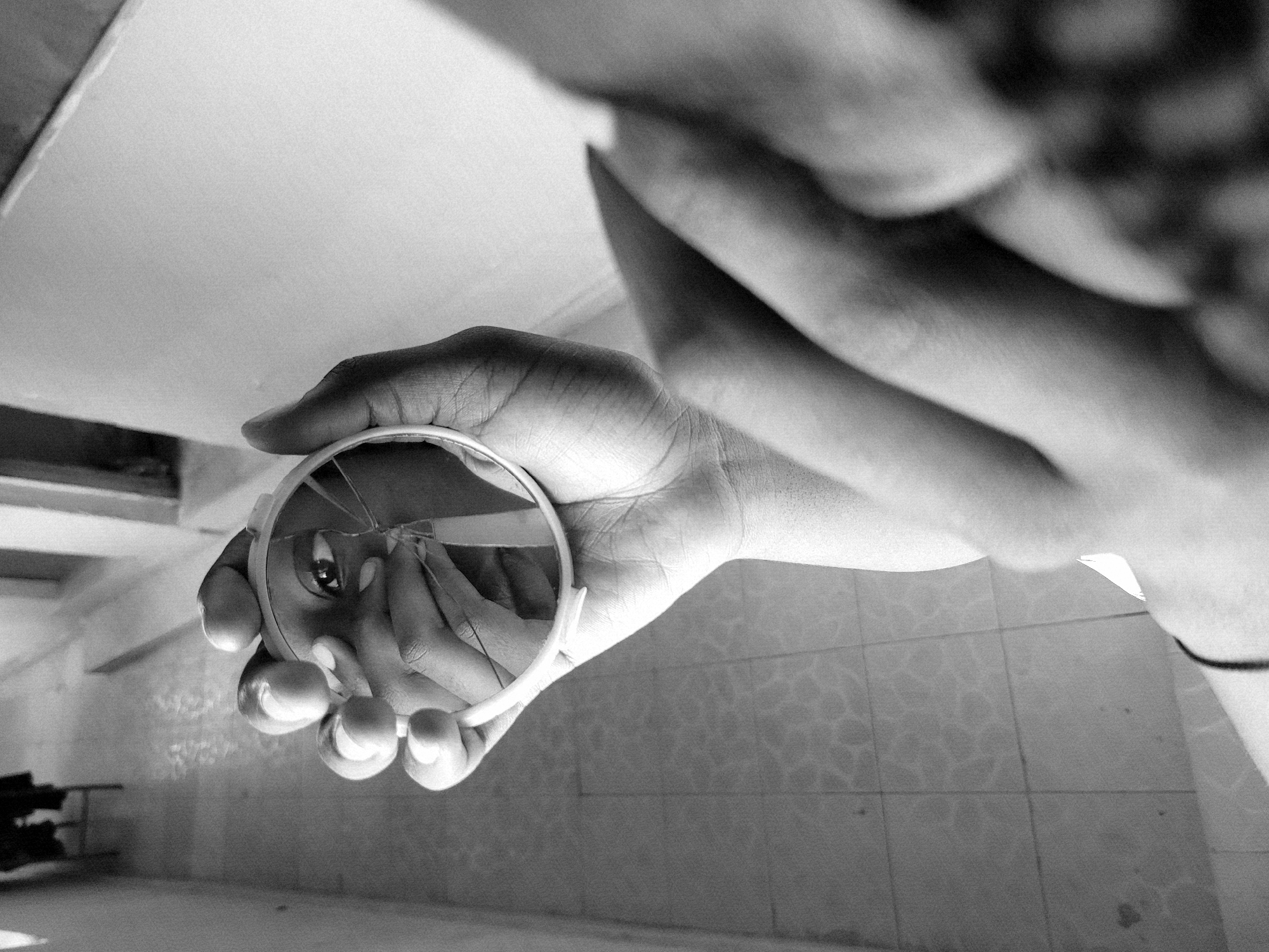 A black and white photograph depicts a person holding a small, round, cracked mirror in their hand. The reflection in the mirror shows a close-up of an eye and fingers manipulating the cracked surface. The person's hand holding the mirror is in focus, while the rest of their arm and torso are out of focus. The background includes a tiled wall and what appears to be the corner of a room. There is no information in the image to establish the location of the city of Jos, Nigeria.