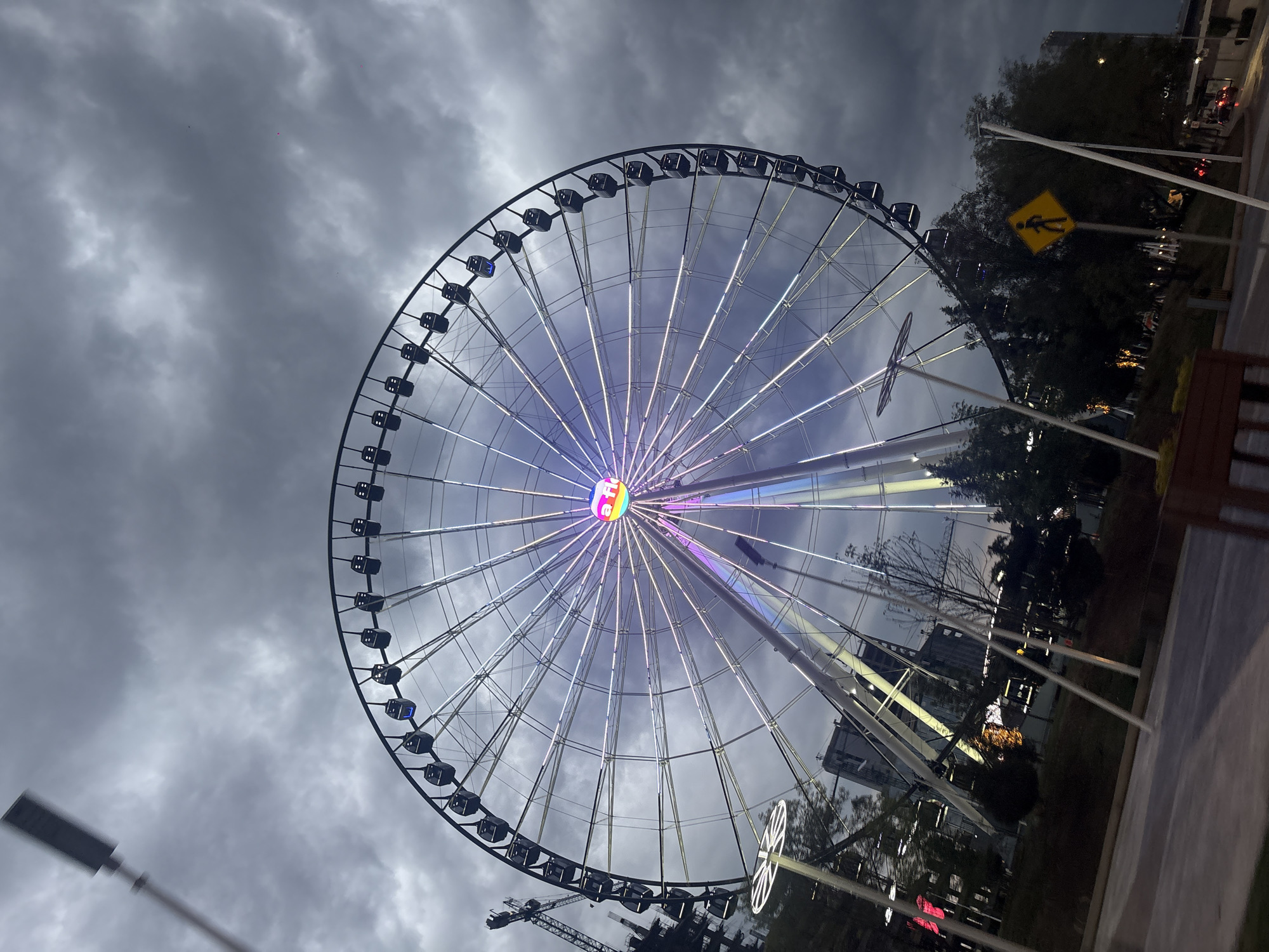 The image shows the "Estrella de Puebla," a giant Ferris wheel located in Puebla, Mexico. The Ferris wheel is prominently featured against a cloudy sky. It is constructed with a black frame and numerous passenger cabins, and its center is illuminated with rainbow lights. The wheel's spokes are also lit up. In the background, there are trees, buildings, and street lights. A pedestrian crossing sign is visible to the right of the Ferris wheel. A construction crane is partially visible at the bottom left of the image.