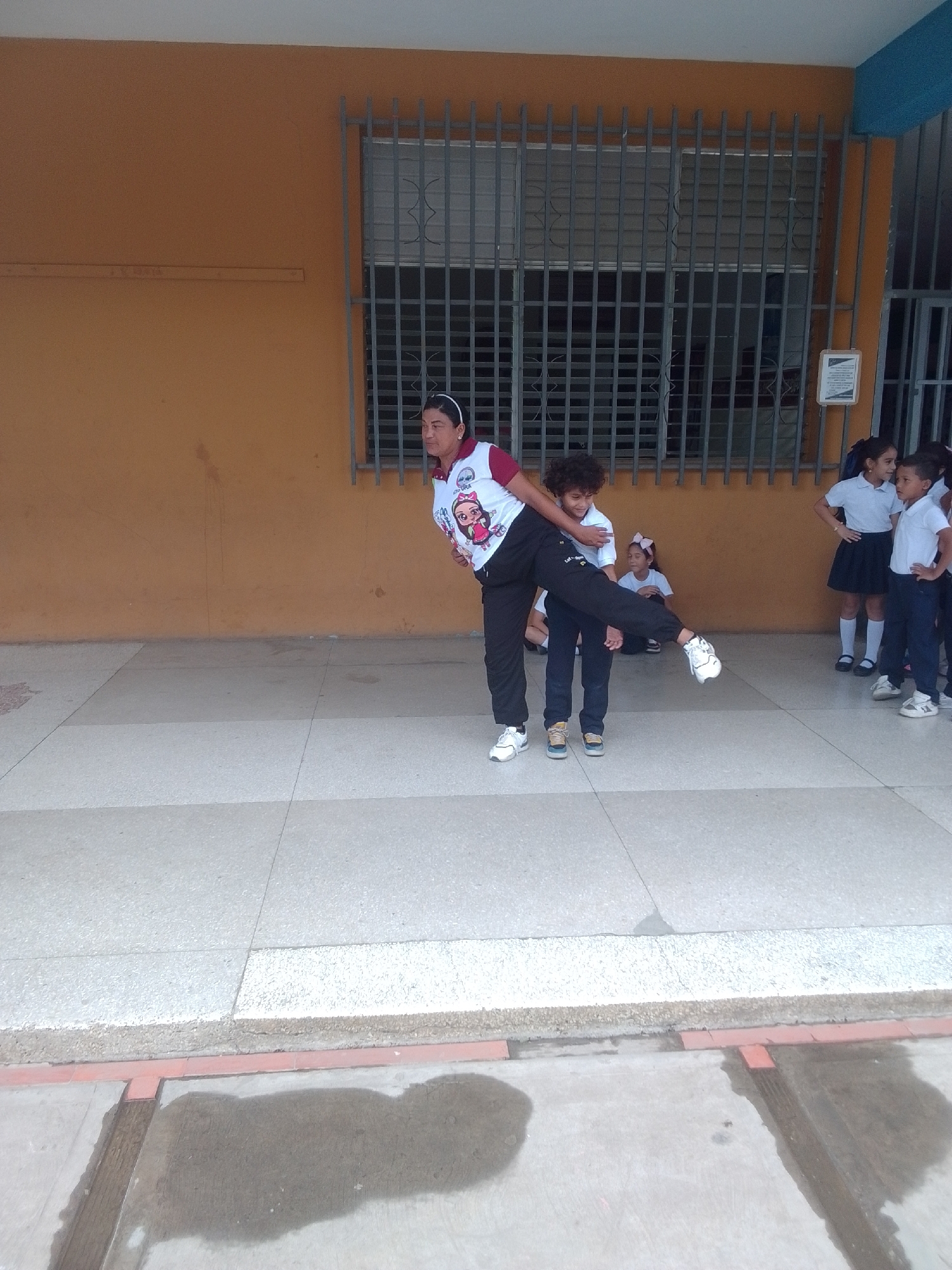 In La Tagua, Venezuela, a group of children in school uniforms are gathered outside a building. The building has an orange facade and a large barred window. Two older children, a boy and a girl, are actively engaged in what appears to be a playful activity. The boy is lifting the girl, who has her legs extended. She is wearing a white shirt with a colorful graphic and dark pants, while he is in a white shirt and dark pants. Several other children, also in school uniforms, are watching. A younger girl with a pink bow in her hair is seated on the ground. The children appear to be in good spirits. The scene suggests a school day or an outdoor school event, likely during daylight hours given the lighting. The ground is paved with a light-colored material, and there are some dark, wet patches, possibly from recent rain or cleaning.