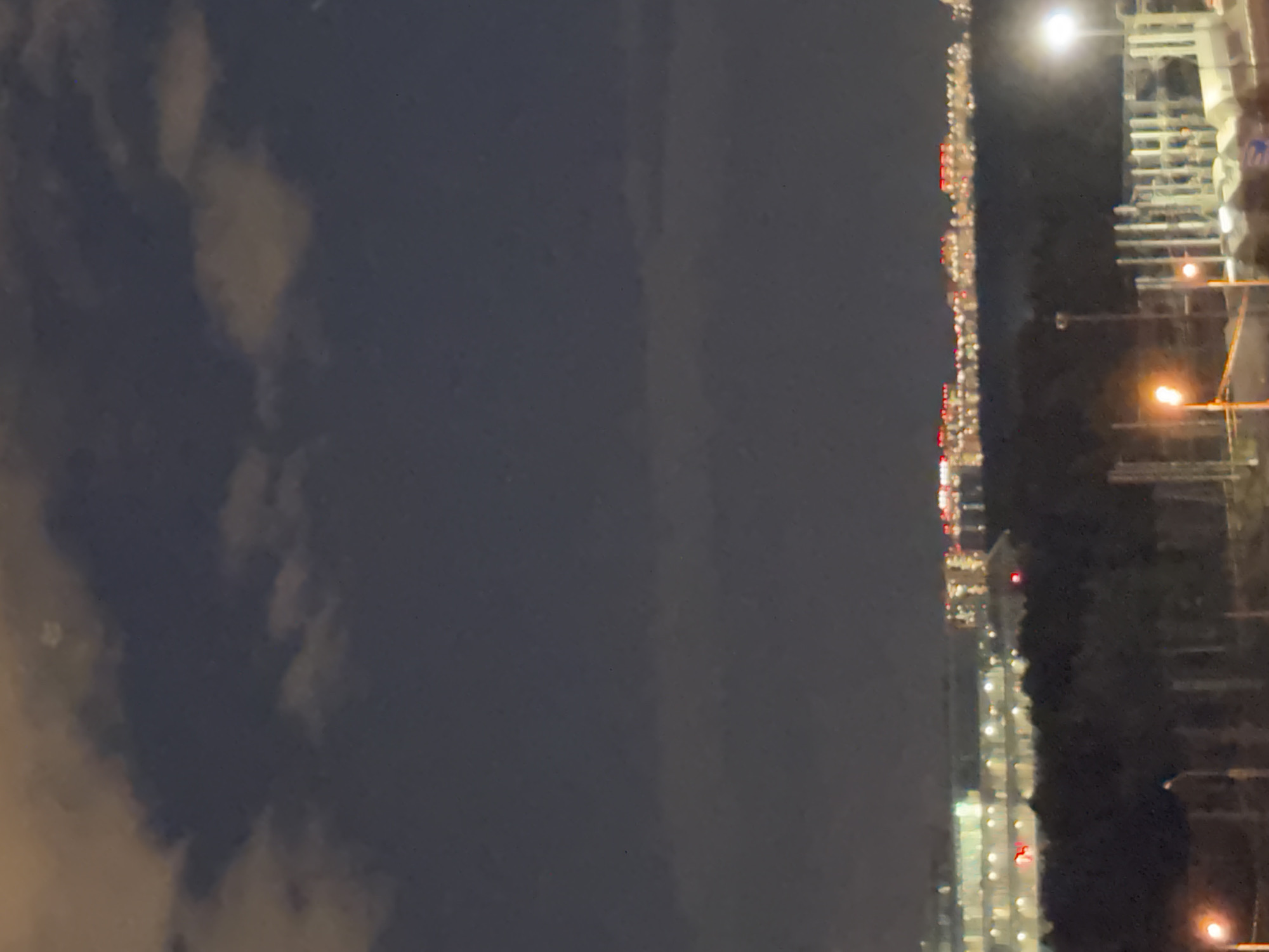 The image is a nighttime photograph taken from an elevated vantage point, likely overlooking a suburban or industrial area. The sky is dark with some clouds, and there are scattered lights in the distance, suggesting a city or town. In the mid-ground, there are trees and structures that appear to be part of a construction site or industrial facility, with some streetlights visible. The far background shows a line of buildings illuminated with many lights, indicating a populated area. Based on the provided context, this scene is located in Odintsovo, Russia.