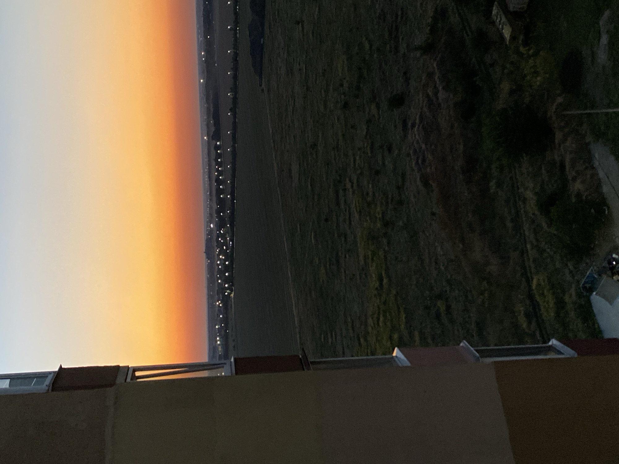 This is an outdoor, high-angle, eye-level shot of a sunset over a rural landscape from a balcony in Chornomorsk, Ukraine. The left side of the frame is dominated by the beige and yellow exterior of a building with windows, a balcony railing, and a portion of a red-painted section. The sky transitions from a pale blue at the top to a vibrant orange and then a deep red towards the horizon. In the middle ground, a dark green field stretches out, interrupted by rows of lights from what appears to be a town or village. The foreground also shows a dark green field with some vegetation and a small, yellow structure on the right. A concrete path leads to a blue object, possibly a small vehicle or cart, at the bottom center of the frame.