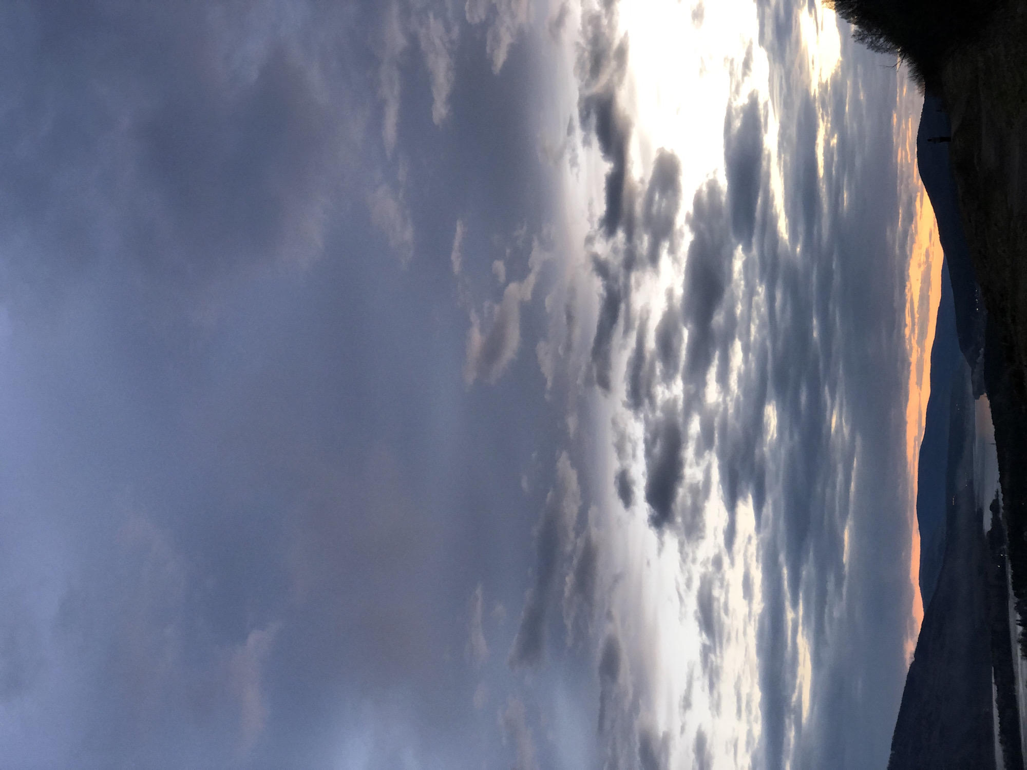 The image presents a landscape under a cloudy sky, likely during sunset or sunrise, given the warm, orange glow illuminating the horizon. The sky is filled with various shades of grey clouds. On the right side of the image, there is a silhouette of a landmass, possibly a mountain range or a large hill, with some greenery visible on the top. Adjacent to the landmass, a body of water is present, reflecting the light.
Given the prompt, the location is likely in or near Krasnoyarsk, Russia, which is known for its landscapes featuring the Yenisei River and surrounding mountains. There are no people directly visible. The interaction involves the natural elements of the sky, land, and water under the specific lighting conditions.