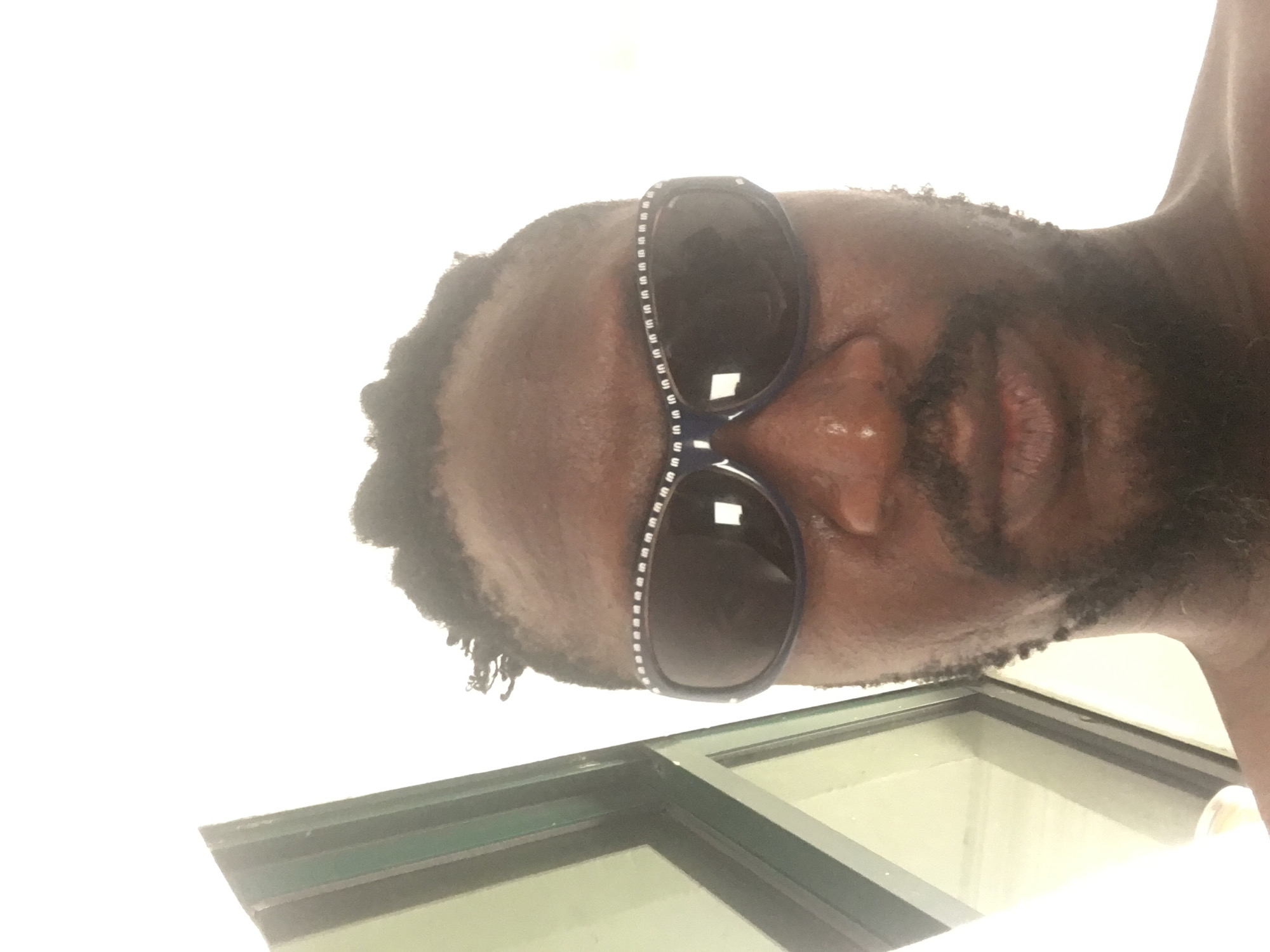 A close-up, low-angle selfie of a dark-skinned male with a beard wearing sunglasses with decorative side panels. He is positioned above a window frame with green trim. The location is not discernible from the image.