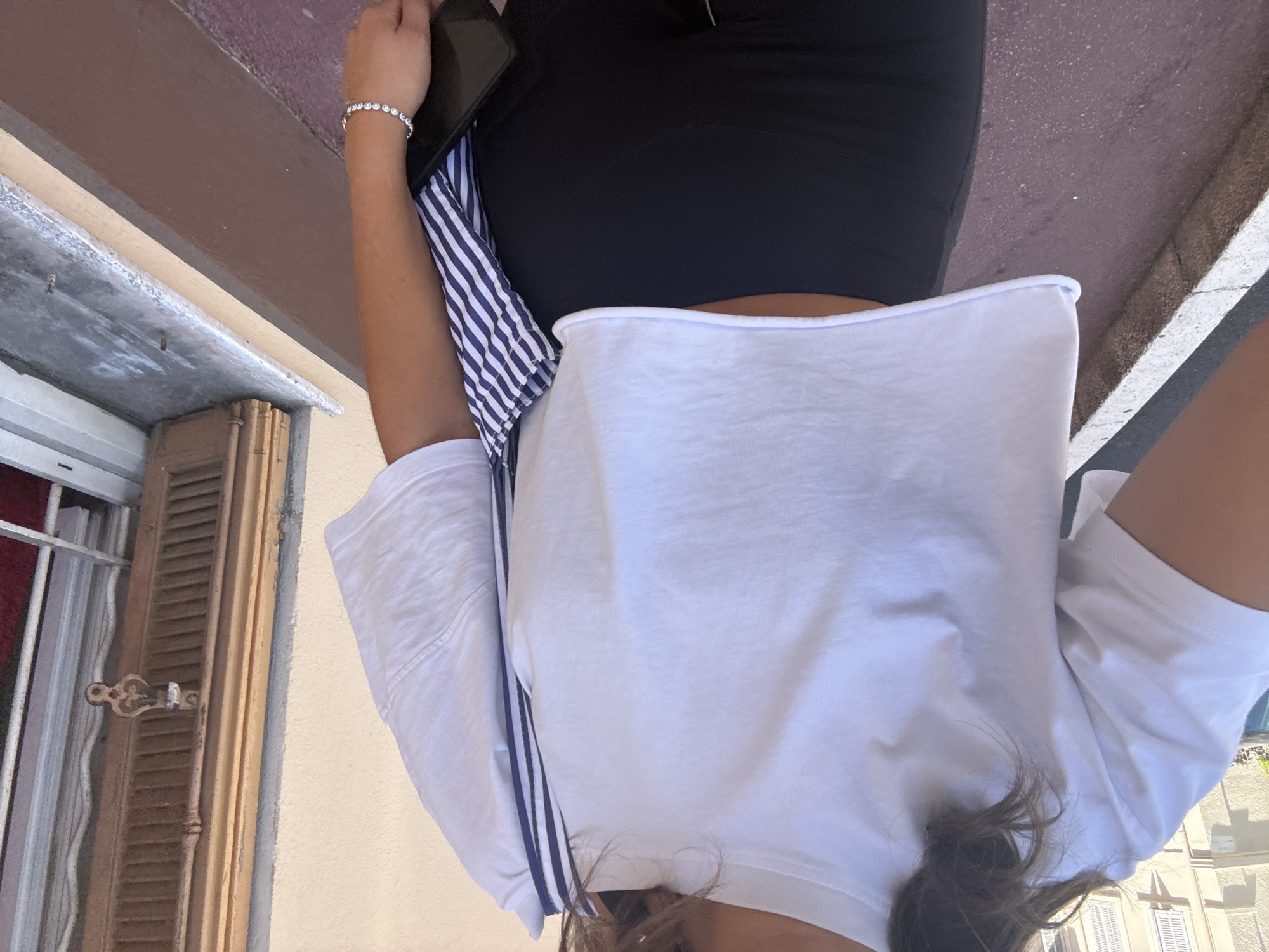 The image shows a person standing on a sidewalk in Cannes, France. The person is wearing a white cropped t-shirt, black leggings, and a striped bag. They are also wearing a diamond bracelet. In the background, there is a building with a window and shutters. The ground is paved with red and gray tiles.