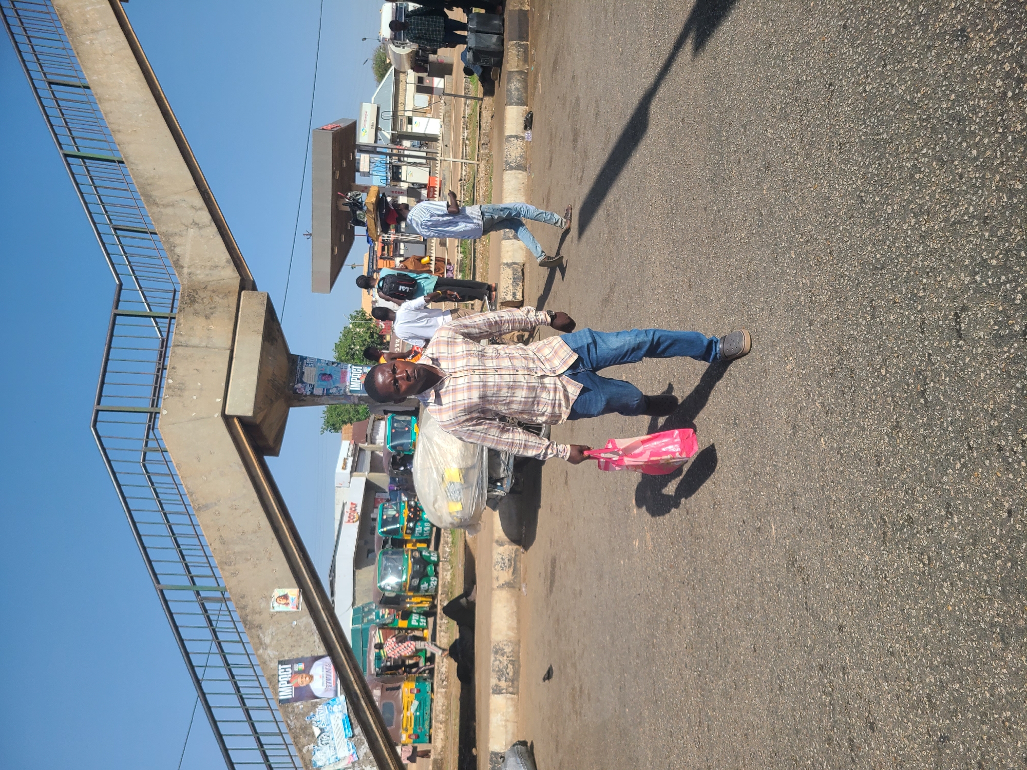 A man is standing on a paved road in Gura Top, Nigeria. He is wearing a plaid shirt and jeans and carrying a plastic bag and a large, clear plastic bag containing items. A pedestrian overpass is on the left side of the frame. Behind the man are other pedestrians and vehicles, including tuk-tuks and cars. A gas station is visible in the background. The sky is blue and clear.