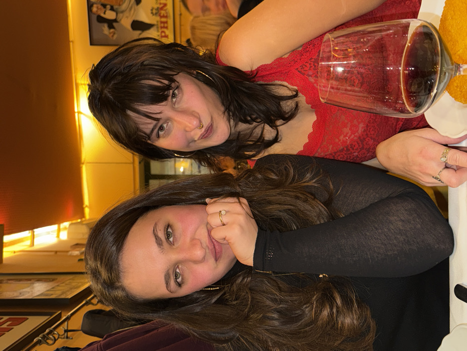 Two women pose indoors at a restaurant. The woman on the left wears a black, long-sleeved top and has wavy brown hair. The woman on the right wears a red, lace top. A glass of red wine and a plate of food sit on the table. A framed image hangs on the wall behind them, and warm lighting illuminates the scene. Both women wear rings on their fingers.