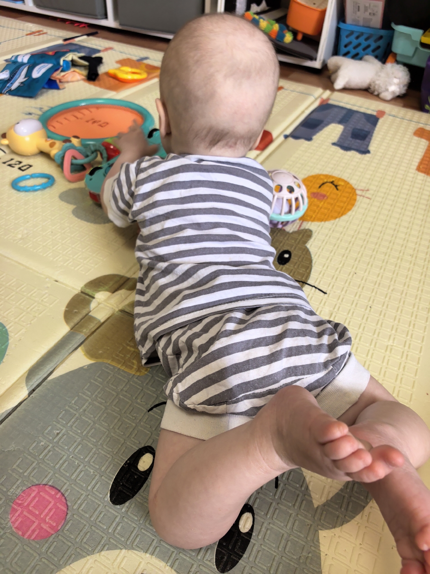 A baby in a striped shirt is prone on a patterned playmat. The baby's head is at the top of the frame and the feet are towards the bottom. The baby is surrounded by toys. The scene appears to be indoors. The location is not discernible from the provided information or image.