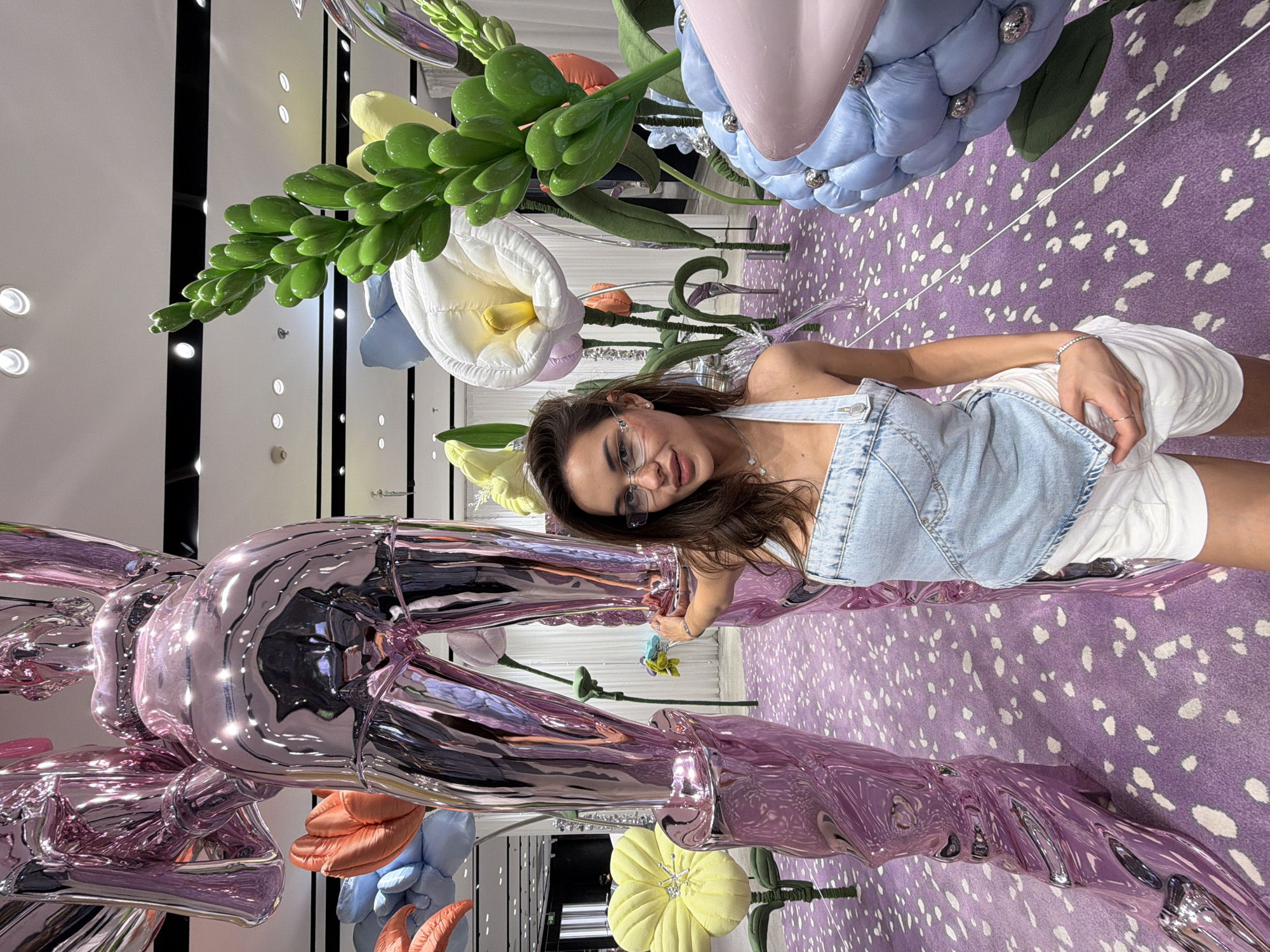 A medium shot shows a woman posing in what appears to be an art installation. The woman has long brown hair and wears glasses, a light blue denim top, and white shorts. She looks towards the camera with a slight smile.
The background consists of oversized, colorful, plush flowers, including white calla lilies, blue and pink flowers, and green foliage. A large, reflective, metallic pink sculpture of a rabbit-like figure is on the left. The floor is covered with a purple carpet with white spots.
The environment appears to be a brightly lit indoor space, possibly a museum or an art gallery. The location is not discernible from the image. It is not possible to confirm the location as Dongcheng District, China from the given image.
