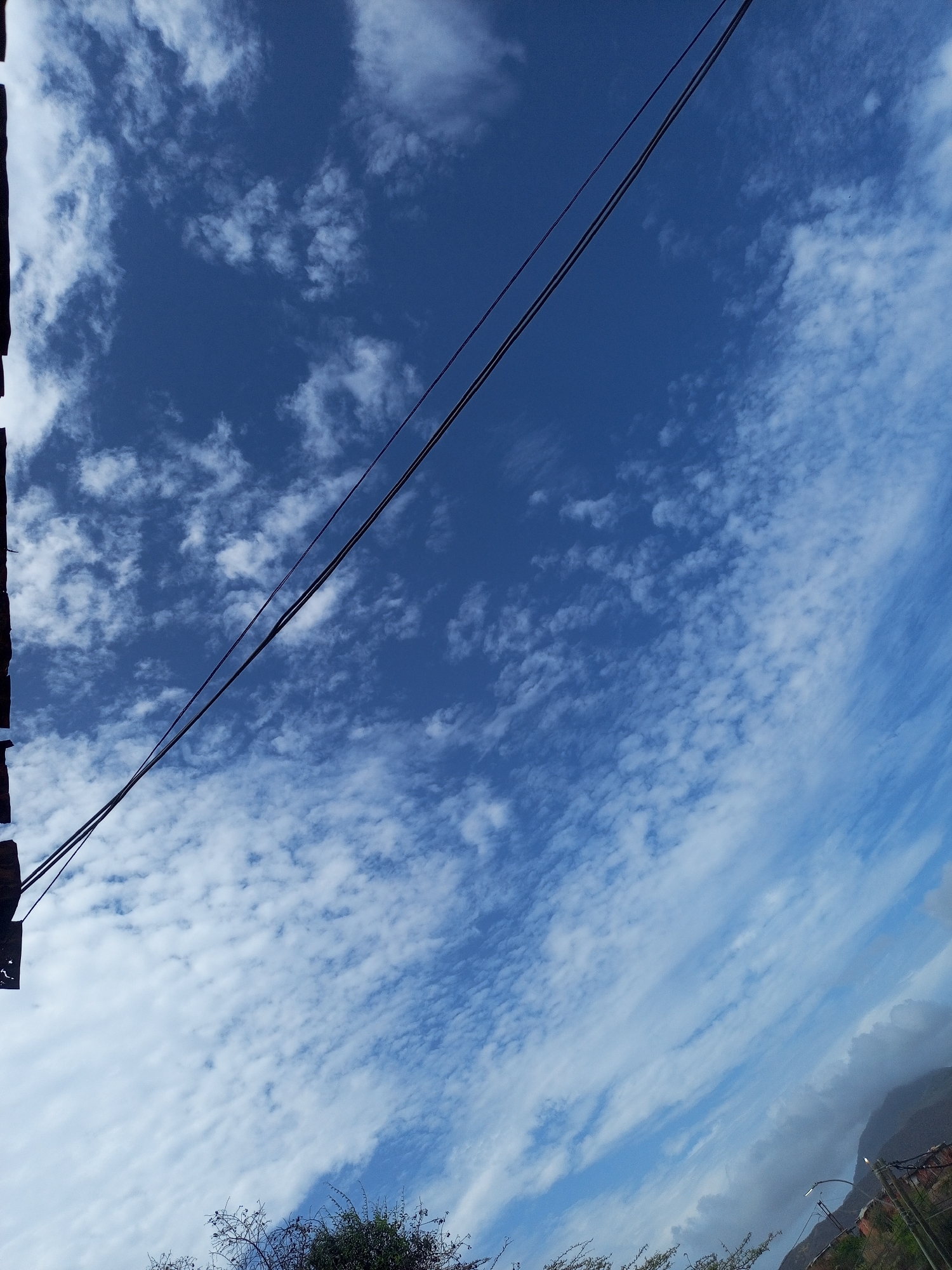 A low-angle, vertical shot captures a vast, deep blue sky filled with scattered white, wispy clouds. Two dark, parallel wires, likely part of a power line, diagonally bisect the frame from the upper left to the lower right. The far left edge of the image is partially obscured by a dark, textured border, possibly a structural element. In the bottom portion of the image, a silhouette of dark foliage, likely trees, spans the width, with a hint of a distant mountain range and a street lamp visible in the lower right corner, suggesting an urban or semi-urban setting near La Tagua, Venezuela. The lighting indicates it is daytime, possibly late afternoon due to the soft illumination and the presence of shadows.