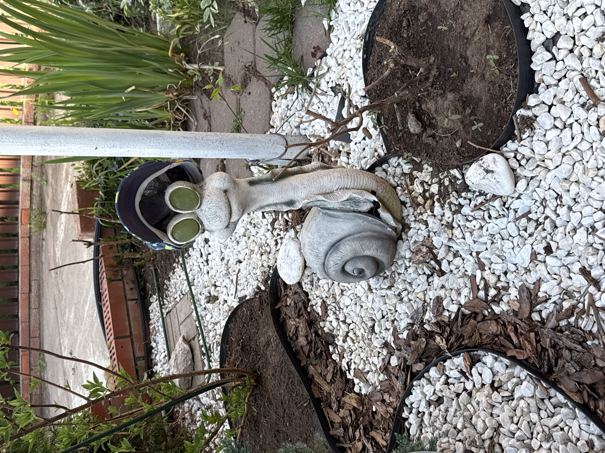 The image depicts a garden scene in Votkinsk, Russia. A decorative snail statue is the central element, lying amongst white gravel. The snail wears a hat and has prominent green eyes. The garden also contains potted plants with soil and wood chips for ground cover. A metal pole is visible, lying parallel to the snail. There are no people visible in the image.