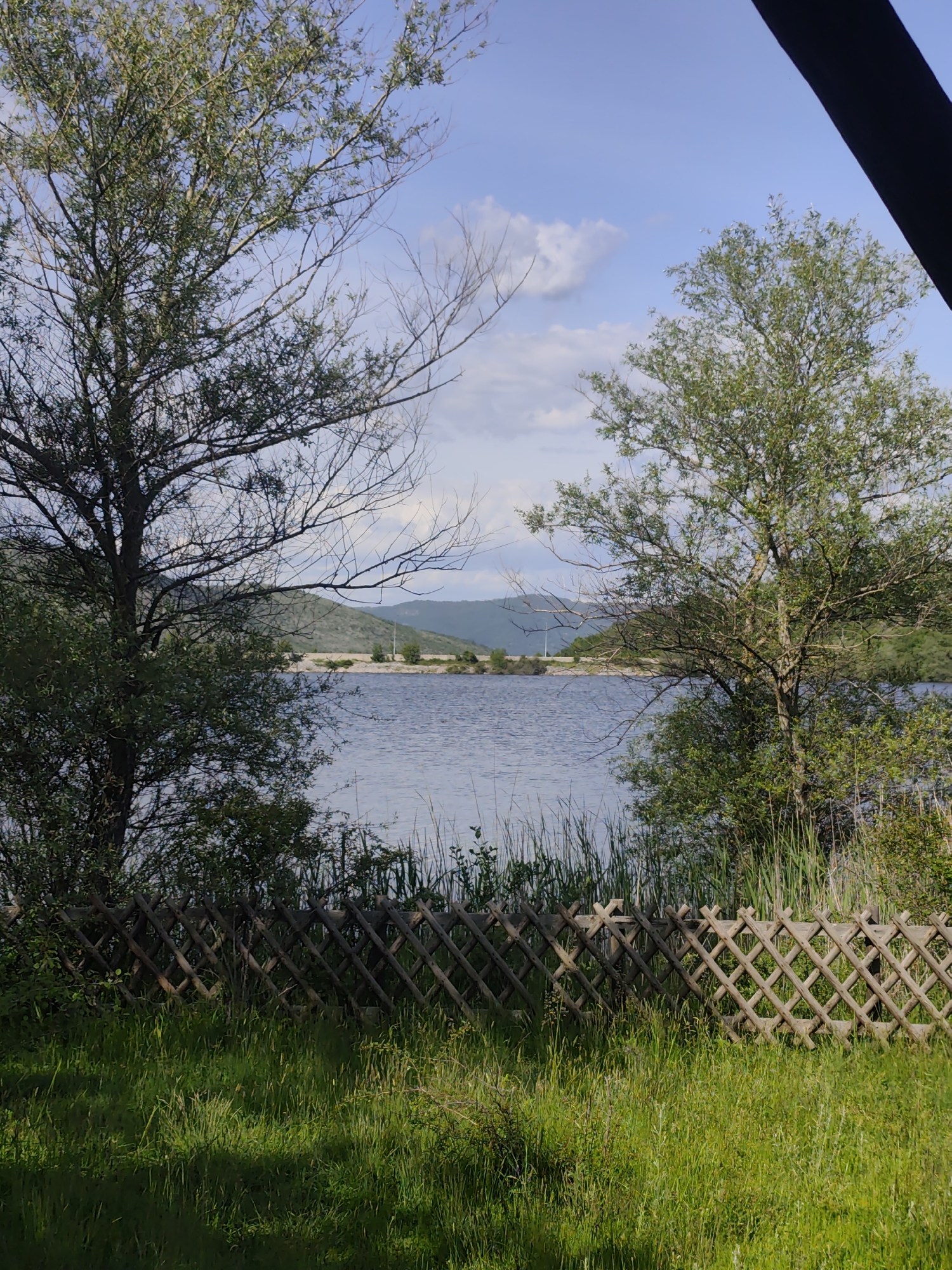 The image depicts a scenic view of a lake framed by trees and a wooden fence. The lake is calm with a shoreline visible in the distance, backed by hills. The sky is light blue with scattered clouds. In the foreground, a wooden lattice fence runs horizontally across the frame, partially obscured by tall green grass. Two trees flank either side of the lake view, with one on the left having sparse branches. The scene is located in the city of Unknown, Unknown.