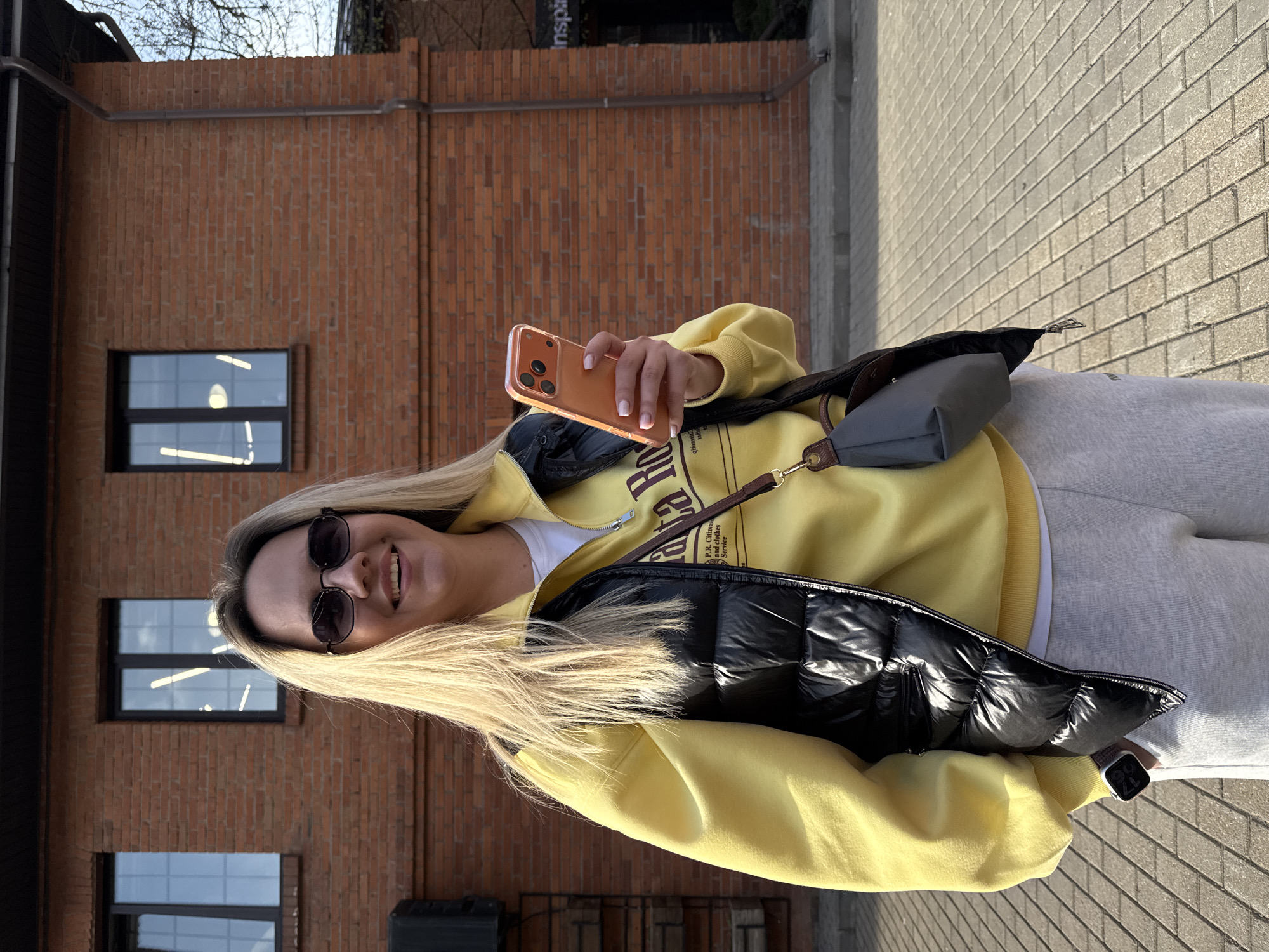 A medium-angle shot shows a blonde woman holding a phone in her right hand with her index finger curled under the phone and her thumb on the screen. She's smiling at the camera, wearing sunglasses, a yellow sweatshirt, a white shirt underneath, a black puffer vest, and grey sweatpants. A grey Longchamp-style purse is slung over her right shoulder. She stands on a sidewalk with grey rectangular pavers. Behind her, a reddish-brown brick building has two visible windows. The location is Moscow, Russia.