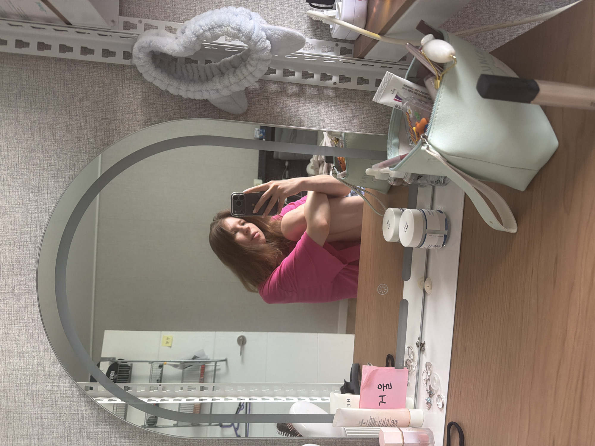 A young woman with brown hair is sitting on the floor in front of a mirror, taking a selfie with her phone. She is wearing a bright pink t-shirt. The mirror reflects a vanity area with various cosmetic items, including makeup, creams, and a makeup bag. A shelf unit is visible to the right, with a white fluffy headband hanging on it. The background appears to be a room, likely a bedroom or dressing area, with textured wallpaper. The image is taken from a slightly elevated angle, looking down at the vanity. It is not possible to determine the specific location in Dongducheon-si, South Korea.