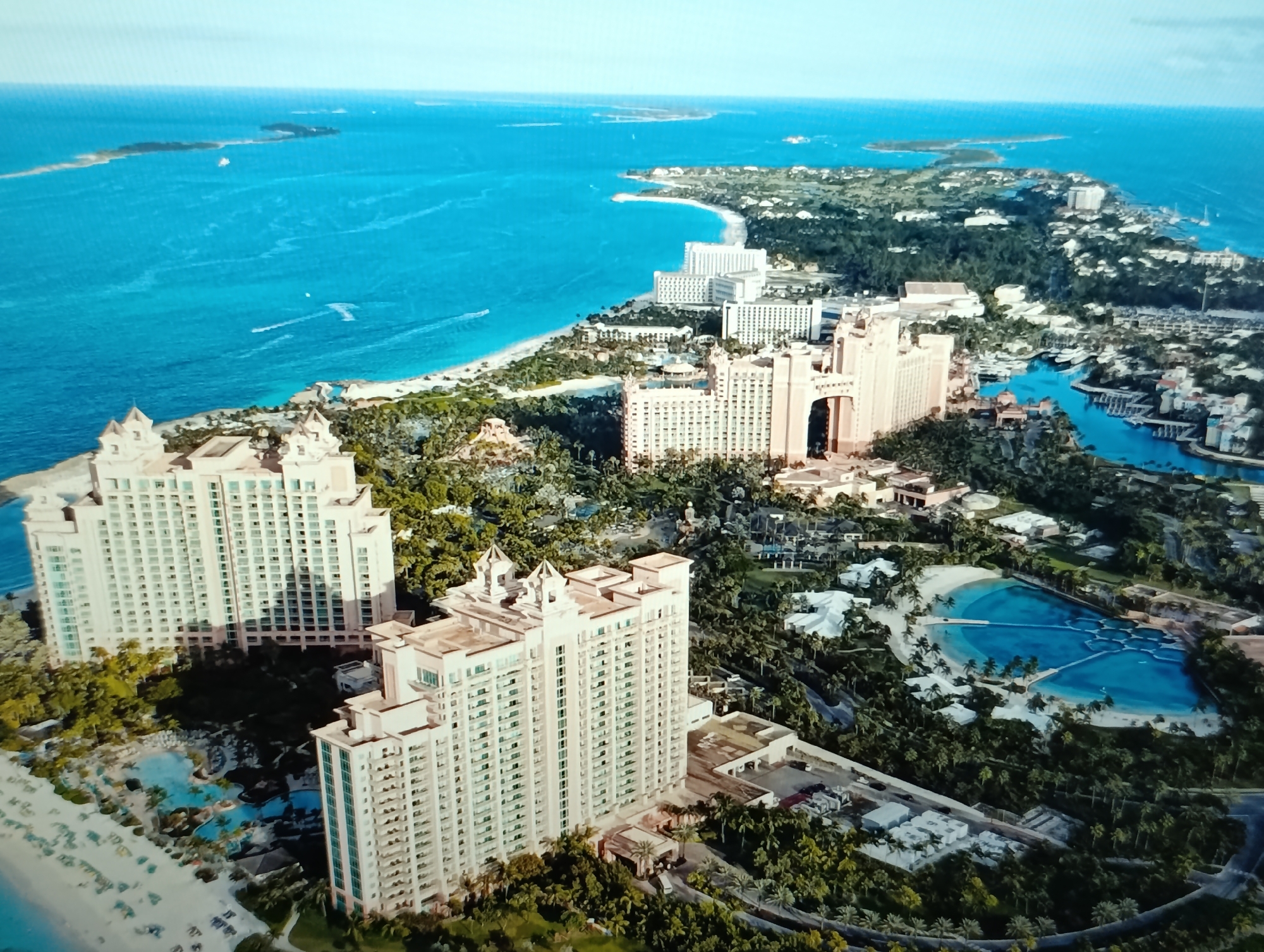 This is an aerial photograph of a resort located on a tropical island. The resort features several large, light-colored hotel buildings, some with distinctive architectural elements like spires. Lush green vegetation, including palm trees, surrounds the buildings and extends towards the coastline. In the foreground, there is a sandy beach and a complex of interconnected turquoise swimming pools, some with water slides. The ocean is a vibrant blue, with a few boats visible on the water and some islands in the distance. The sky is clear and pale blue. The location is not in the city of Dubăsari town council, Moldova.