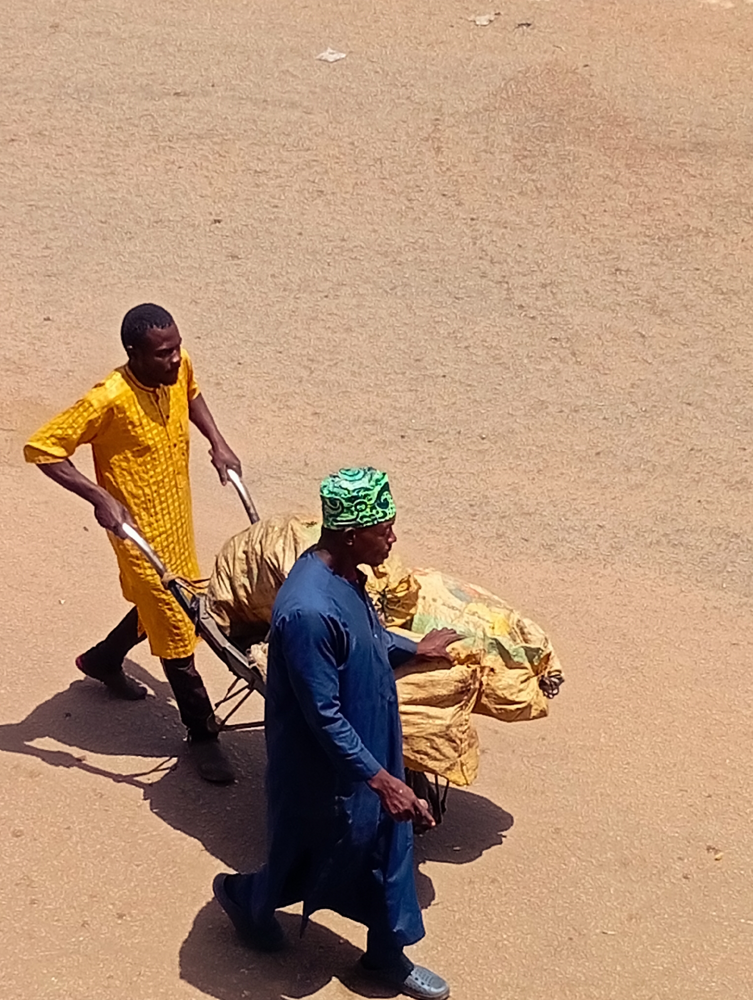 Two men are pushing a handcart filled with large sacks. The man in the foreground is wearing a dark blue robe and a green, patterned headwrap. He is pushing the cart with his left hand. The man behind him is wearing a yellow robe with a patterned texture and black pants. He is gripping the handles of the cart with both hands. The handcart has metal handles and a dark frame. The sacks on the cart are light brown and appear to be made of a woven material. They are large and irregularly shaped. The ground is a dusty, light brown color, suggesting it is an outdoor, possibly unpaved, surface. The scene appears to be set in Bukuru, Nigeria, under a bright sun, as indicated by the long shadows cast by the men and the cart. The lighting suggests it is daytime, likely mid-morning or afternoon.
