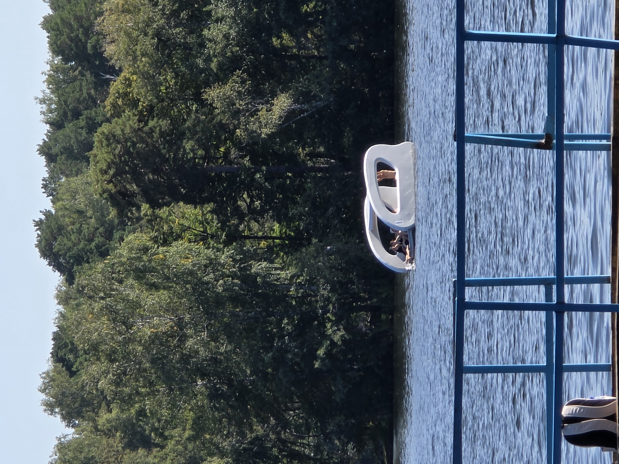 This is a medium shot, taken outdoors on a sunny day, of a peculiar white pedal boat on the water. The boat has a futuristic, rounded design with a partial canopy, and two people are visible inside, seated and relaxed. The boat is on a body of water, likely a lake, with a dense forest of green trees forming the background. In the foreground, a blue metal railing is visible, and a backpack rests on a wooden surface in front of it. The scene suggests a leisurely outing on a calm day. This scene is on the shore of Lake Chebarkul in the city of Chebarkul, Russia.