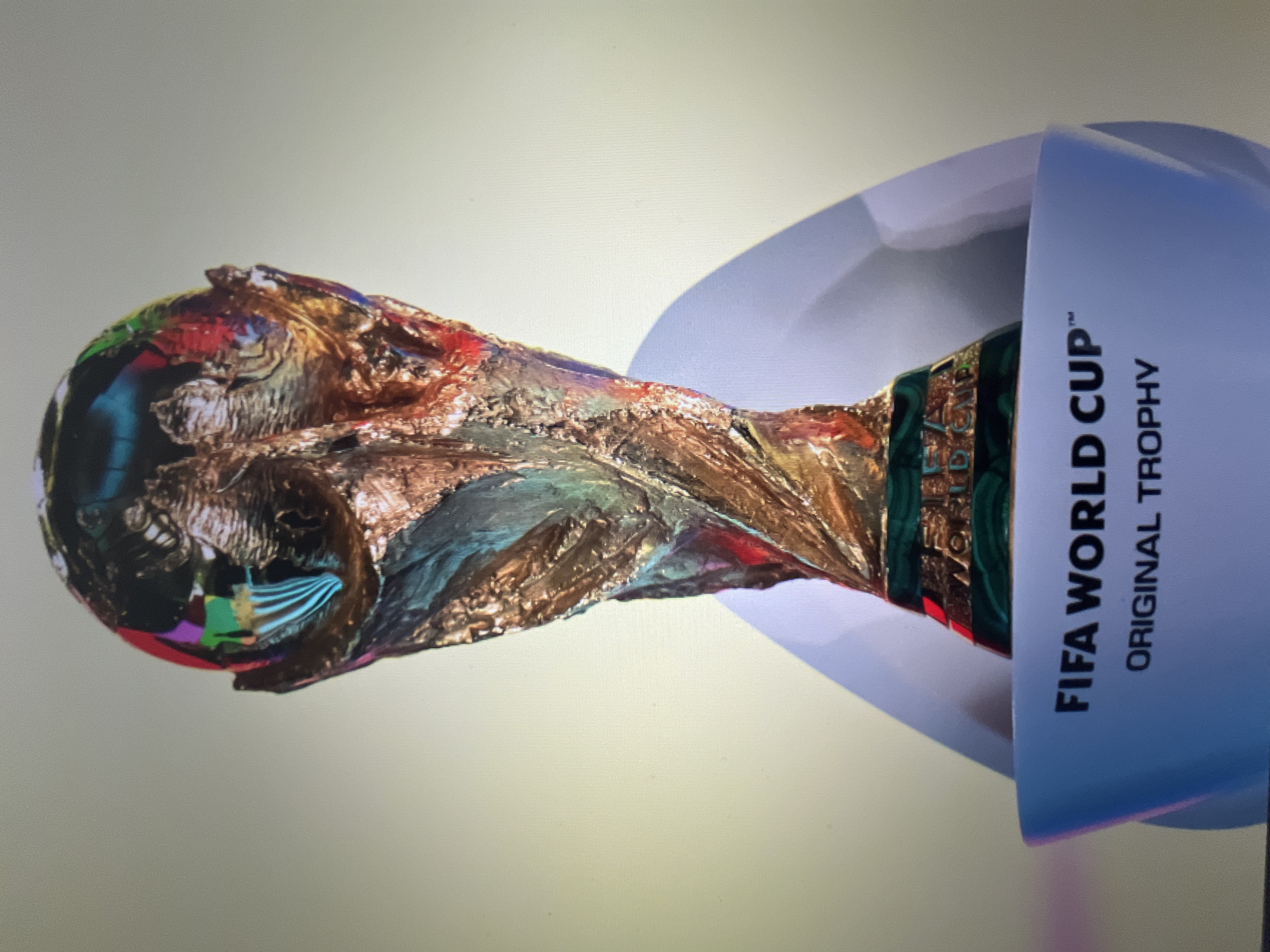 The image displays the FIFA World Cup trophy, positioned on its side, against a plain, light grey background. The trophy is made of a reflective golden material, with a highly textured and intricate surface that shows various colorful reflections, including reds, blues, greens, and purples, particularly on its upper, spherical part. The lower part of the trophy's base features visible green and gold details.
The trophy is partially enclosed within a white, curved display case or stand. On the side of this white stand, prominent black capital lettering reads: "FIFA WORLD CUP™ ORIGINAL TROPHY". The overall lighting appears soft and even, suggesting an indoor setting. There is no specific activity depicted, but rather a static display of the iconic football award. The image exhibits characteristics of being a photograph taken of a digital screen, with subtle pixelation visible across the surface. While the location context of Varna, Bulgaria, was provided, there are no discernible visual cues within the image itself to confirm this location.