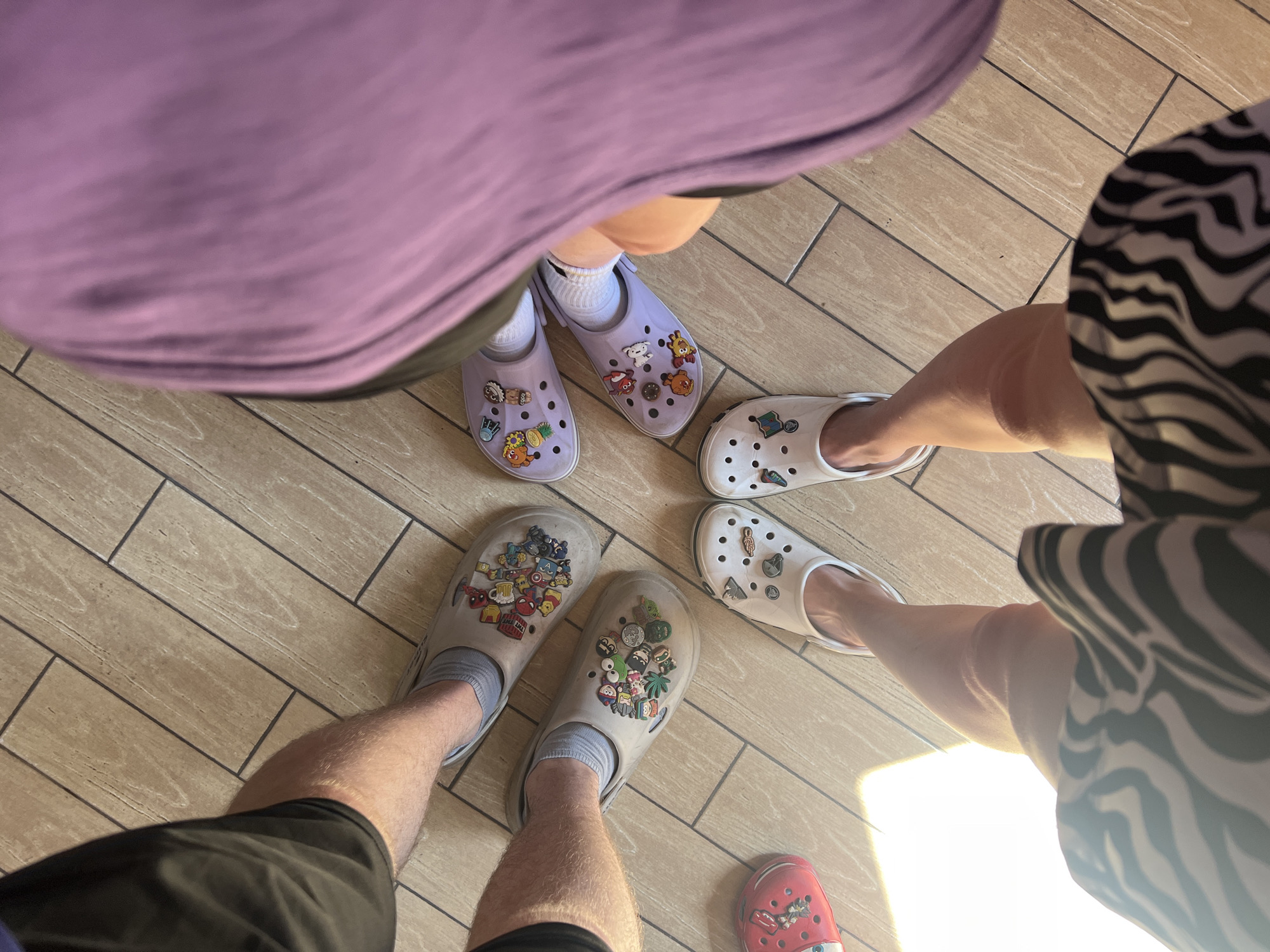 Four individuals stand in a circle on a tiled floor. One person is wearing a purple shirt and lavender Crocs adorned with cartoon charms. Another person is wearing a zebra-striped top and white Crocs. A third person is wearing black shorts and gray Crocs featuring superhero and cartoon charms. A fourth person's lower legs and red Crocs with cartoon charms are partially visible. The floor is light brown with a wood-grain pattern. A patch of bright sunlight is visible near the bottom right. The location is not known to be Grishanki, Russia.