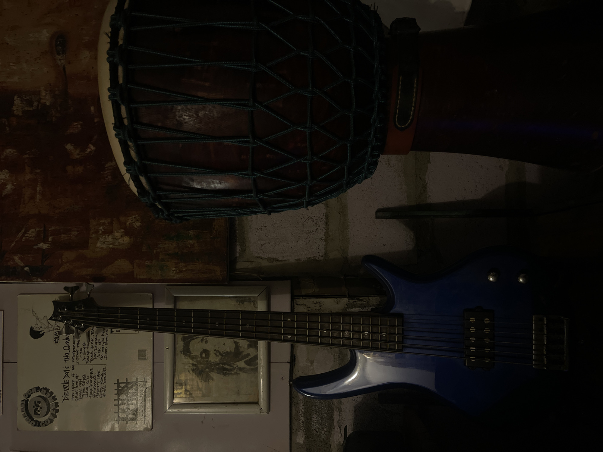 This is a low-light, indoor photograph of a blue electric bass guitar and a djembe drum. The guitar is positioned horizontally in the lower half of the frame, with its neck and frets clearly visible. The djembe drum is situated above the guitar, its dark wooden body and netting also discernible.
On the wall behind the guitar, there appears to be a framed picture or poster with some text and an image of a person. The visible text is primarily in English and seems to be related to music or an album, with song titles and dates like "DIERRE DON & THA DOVE," "1995," and "Street Life." The background wall has a rough, textured, and somewhat aged appearance.
The overall mood is dark and intimate, possibly suggesting a music studio, rehearsal space, or a personal collection of musical instruments. The lighting is dim, with some ambient light illuminating the objects. There are no people visible in the image. The location context provided is Chalong, Thailand.