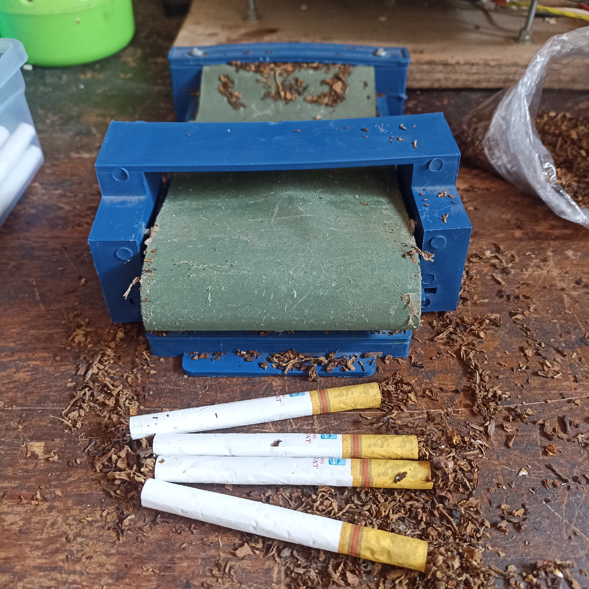 The image depicts a close-up, eye-level view of a wooden surface with scattered loose tobacco and several partially rolled cigarettes. A blue cigarette-rolling machine is centrally positioned, containing some dried tobacco leaves. To the left of the machine, a clear plastic container holds several white cigarette filters. To the right, a clear plastic bag is filled with loose tobacco. Four finished cigarettes are arranged in a slight diagonal line in the foreground, lying on top of the scattered tobacco. The overall scene suggests a setting where cigarettes are being manually rolled. The image's lighting is natural, casting soft shadows, and the background is blurred, drawing focus to the foreground objects. The location is identified as Magelang, Indonesia.