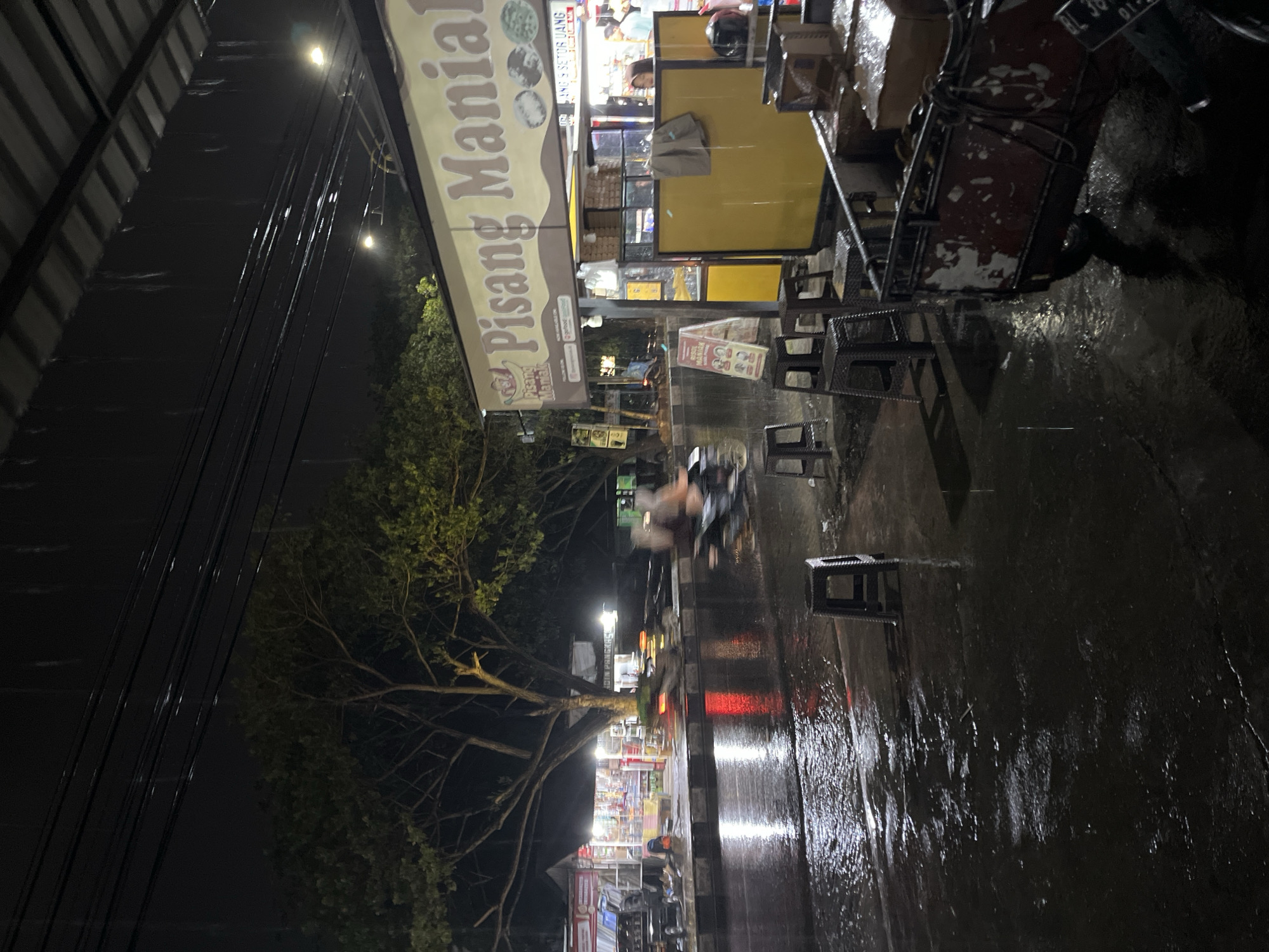 This is a nighttime, low-angle shot of a street scene in Banda Aceh, Indonesia during a rain shower. The foreground is dominated by a wet, reflective street, indicating recent or ongoing rainfall. Several plastic stools are placed on the wet pavement. To the right, a red and white utility cart, loaded with boxes, is parked, with a motorcycle visible to its right.
In the midground, a yellow food stall with a sign reading "Pisang Maniak" (Banana Mania) is illuminated, with people visible inside. A menu board stands in front of the stall. Further down the street, there are other illuminated stalls and shops, with a large tree providing shade. Traffic is present on the street, with a motorcycle in motion creating a blur, suggesting a moderately busy thoroughfare. The overall atmosphere is that of a wet, urban night market or food street.