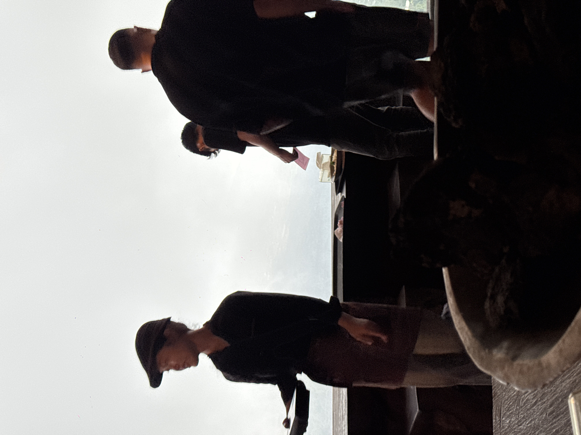 In Kintamani, Indonesia, three individuals are visible inside a dimly lit establishment with a large window overlooking a hazy outdoor environment. A person wearing a dark cap and dark top stands on the left, holding a dark rectangular object. To the right, another individual in a dark top faces away from the camera. The third person, also in dark attire, is partially visible on the far right. The interior features a dark counter or table in the foreground and background.