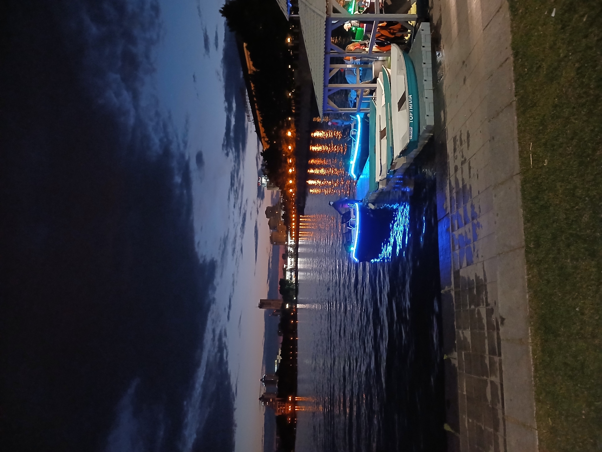 At dusk, a waterfront scene unfolds in Yekaterinburg, Russia. A dark, cloudy sky looms overhead, with scattered lights from a distant city skyline reflected in the calm water. A paved promenade runs along the edge of the water, bordered by a strip of grass. A covered dock area features several small boats, some with blue neon lights. A person stands on the dock near a boat, and several life vests are visible hanging on a structure. Another boat, also illuminated with blue lights, is partially submerged in the water next to the dock.