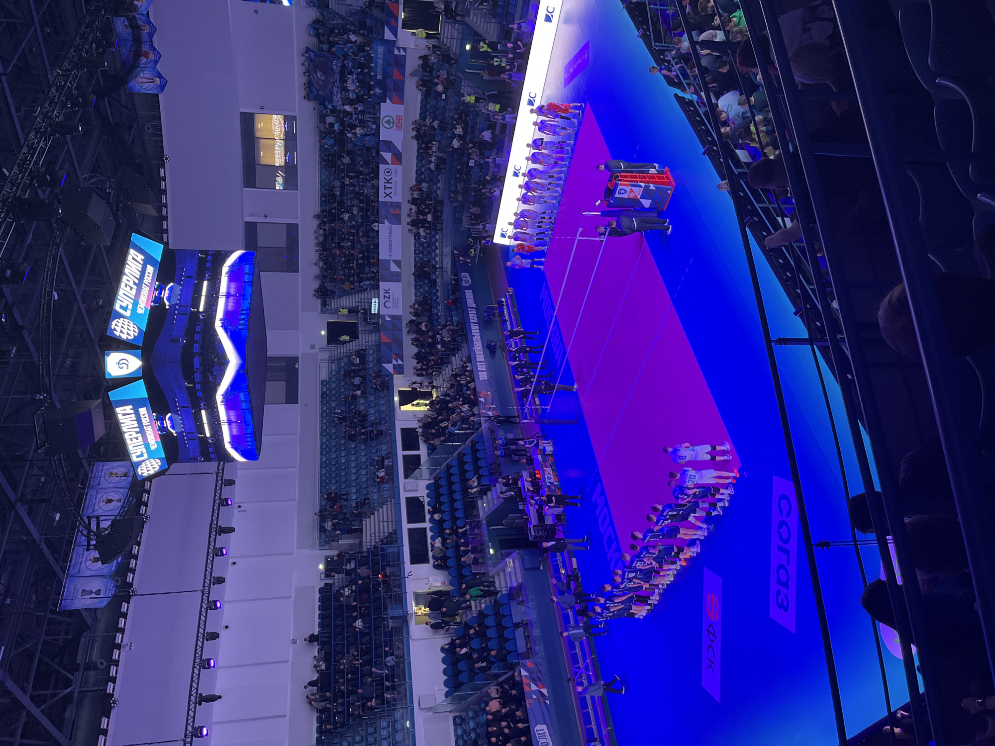 The image shows an indoor volleyball game being held in Moscow, Russia. The image shows players lined up on either side of the volleyball court, likely for a ceremony or the start of a match. Large screens display the name of the "Super League Championship of Russia." The venue appears to be filled with spectators in stadium-style seating. There are advertisements visible, including logos for "ФСК," "согаз," "OZK," "НОВОТРАНС," and "XTKO РЕСУРС ОЙЛ." "ПИТЕРСКИЙ ПРАЙД" is also visible.