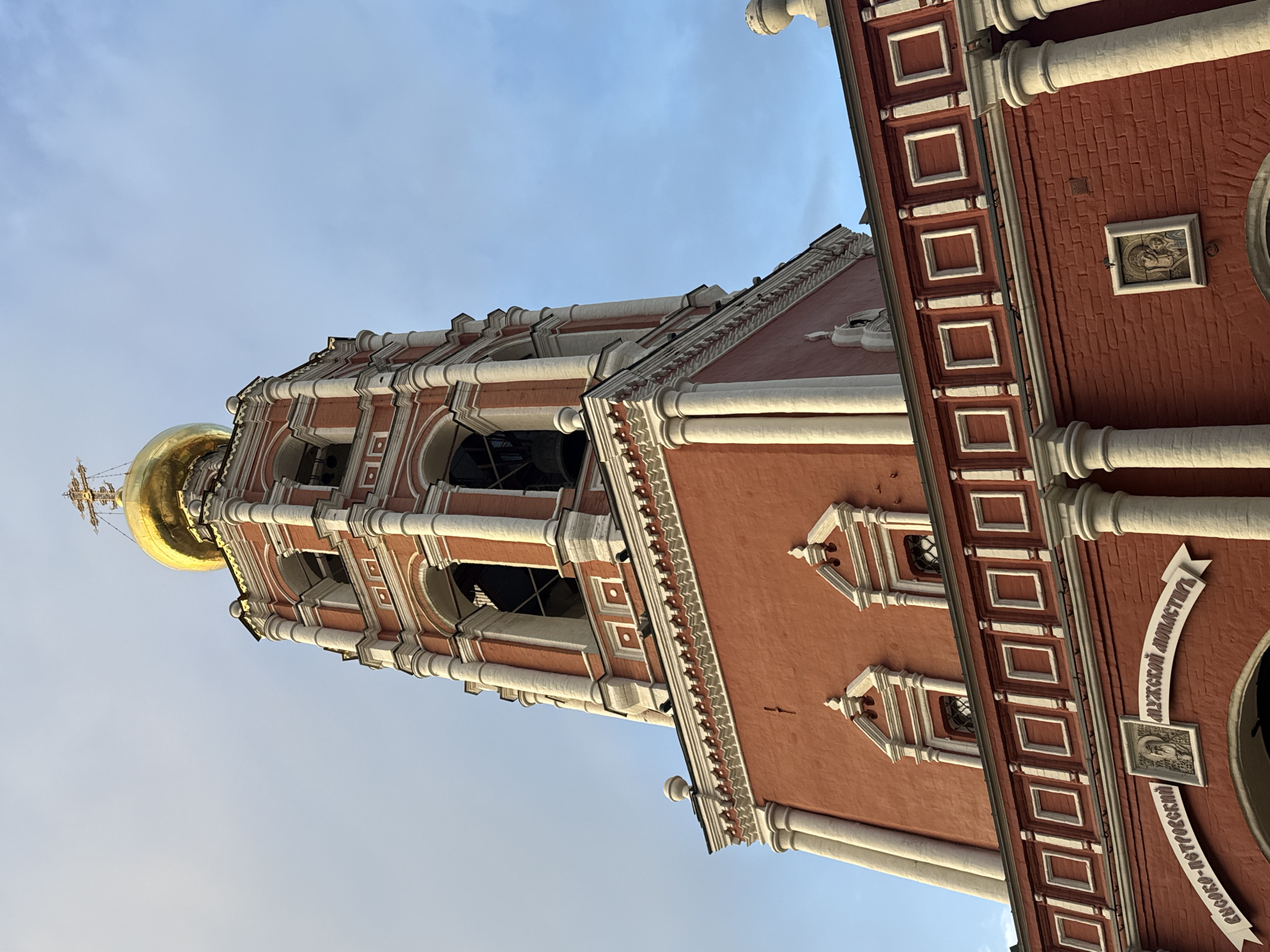 The image shows a view looking up at a red and white building with a gold dome, identified as part of the Vysoko-Petrovsky Monastery in Moscow, Russia. The architecture features tiered structures with white columns and arches, ornate detailing, and small windows. The sky is light blue.
