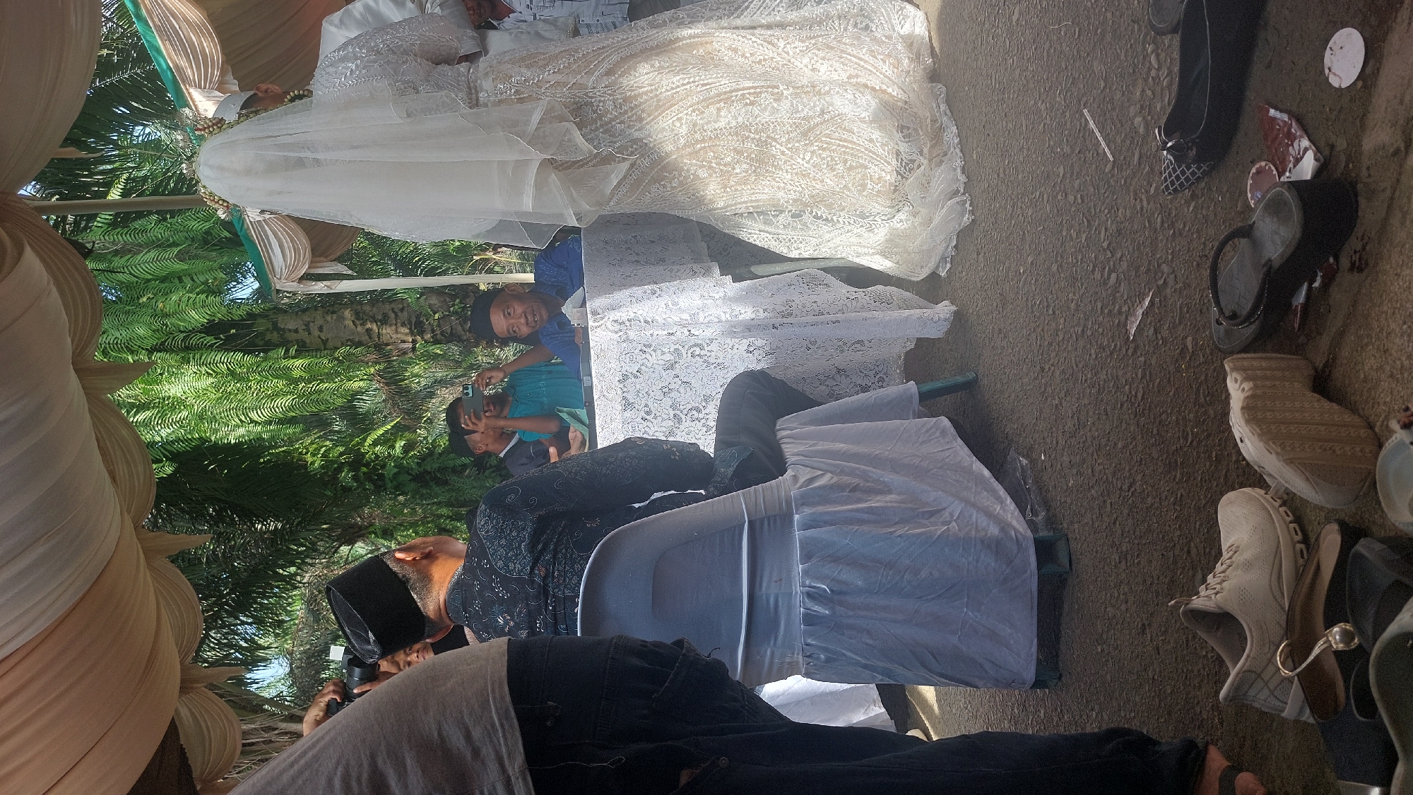 This is a medium shot photograph taken outdoors under a tent, with lush green foliage visible in the background, suggestive of a tropical or subtropical environment. The ground is paved with asphalt. The scene appears to be a wedding ceremony.
In the foreground, a person's legs and lower torso are visible on the left, dressed in dark blue jeans. Scattered on the ground are various footwear items, including a light-colored athletic shoe, a pair of dark flip-flops, and other dark shoes.
In the mid-ground, a man wearing a black peci (a type of cap) and a black patterned shirt is seated in a white chair covered with a white cloth. He is looking away from the camera. To his right, a table draped with a white lace tablecloth is partially visible. Behind the table, a man in a blue shirt and a dark peci is seated and looking towards the camera, appearing to be engaged in conversation or observation. Two other individuals are visible behind him, one holding a mobile phone as if taking a picture.
On the right side of the frame, a bride is visible, her face obscured by a white veil. She is wearing an elaborate white wedding dress with intricate lace detailing. She is standing and appears to be facing away from the viewer, possibly towards the groom, who is not clearly visible but implied to be on her side. A man in a white shirt is standing next to the bride.
The overall impression is a candid moment captured during an outdoor wedding celebration in a warm climate. The location is identified as Sigara Gara, Indonesia.