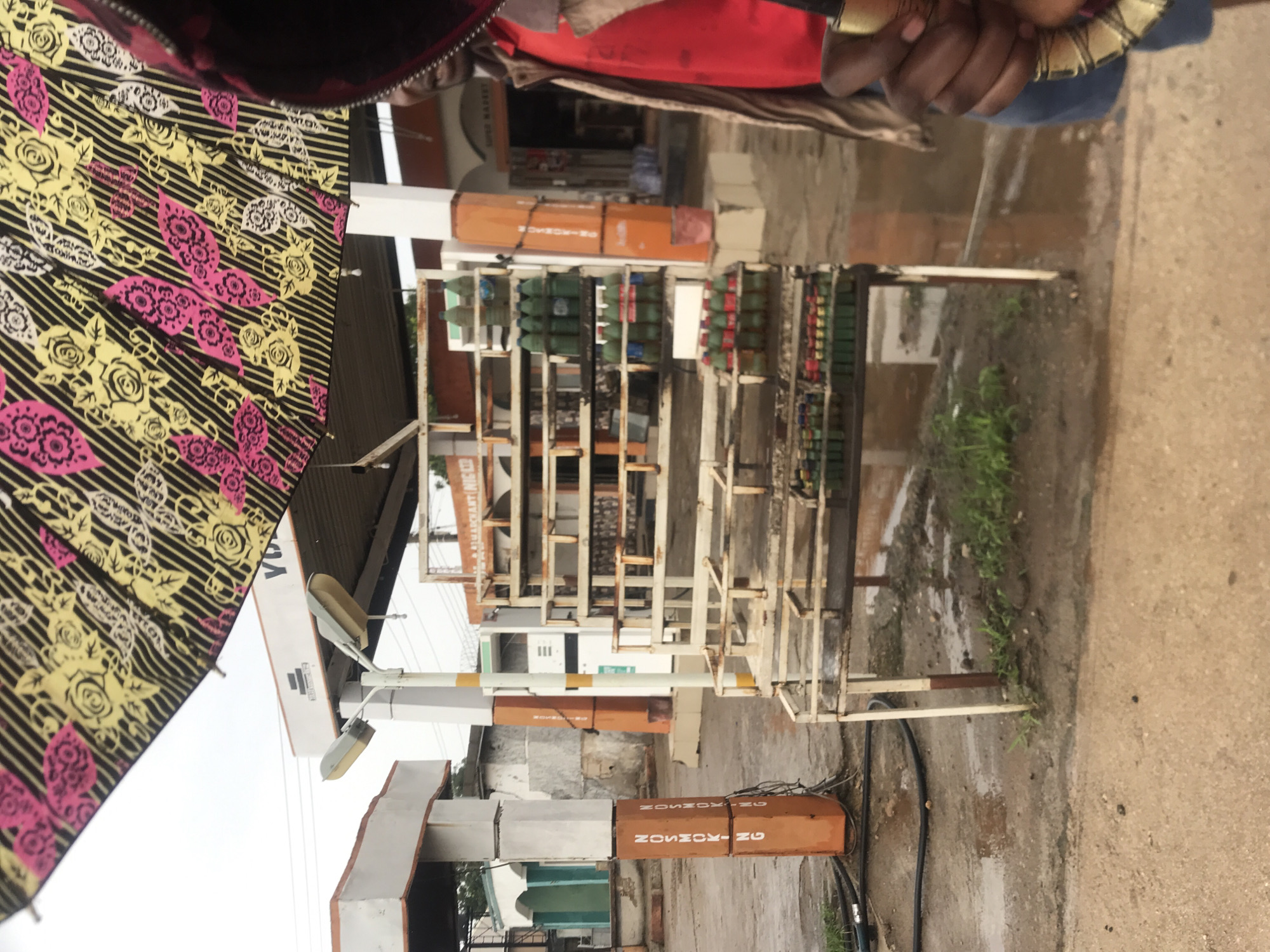 This image, oriented correctly, depicts an outdoor commercial scene in Rafin Par, Nigeria, featuring what appears to be a gas sales or distribution point. In the foreground, the upper torso and hands of a person are partially visible on the right, holding a black umbrella patterned with yellow and pink floral and striped designs on the left. The ground is damp and muddy in places, suggesting recent rainfall.
The central part of the image is dominated by multiple white metal shelving units, organized in rows. These shelves are filled with numerous small gas cylinders or bottles, predominantly green and red, indicating a retail or refilling operation for cooking gas. Behind these units, an orange, vertical cylindrical dispenser or storage tank bears the white, stenciled text "NOSMOSON-NG." A second, similar orange structure is visible further to the left.
In the background, under an overhang, a sign with "SUPER MARKET" is partially visible. Another white sign shows fragmented text, including "VO." Further back, part of a sign with "ALHAJI AKM" can be seen. White poles supporting street lights are also present. The sky appears overcast, suggesting daytime. The overall setup portrays an informal yet structured outdoor retail environment common in the region.