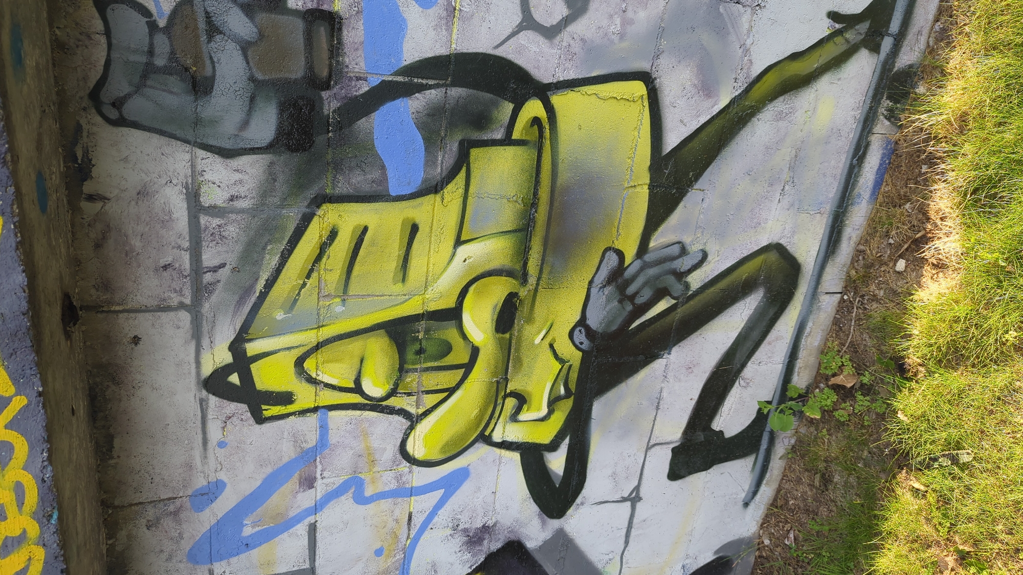The image shows graffiti art on a concrete wall. The graffiti depicts a cartoon-like character in yellow with a black outline, featuring large eyes and a long nose, with thin, black, spindly legs and arms. A small, grey, humanoid hand is depicted in the lower right section of the character's body. Behind the yellow character, there are abstract blue and grey spray-painted shapes, and above it, there are yellow markings on a concrete ledge. The wall appears to be part of an outdoor structure, possibly an underpass or bridge, with grass and dirt at its base. The lighting suggests it is daytime. The graffiti style is typical of urban street art. Without further context, the specific location in Montreal, Canada, cannot be determined.