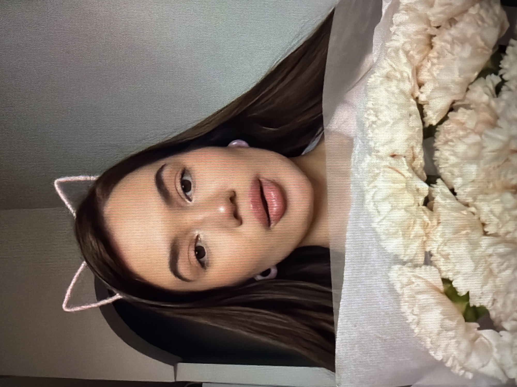 In this image, a young woman with long brown hair and a pink cat-ear headband is holding a bouquet of light-colored flowers. She is wearing light pink hoop earrings. The background appears to be an indoor setting with textured grey walls and a dark archway. The lighting is soft, and the image quality suggests it might be a screenshot from a video call or a lower-resolution photo. The location in Sirius, Russia is not discernible from the visual elements of the image.
